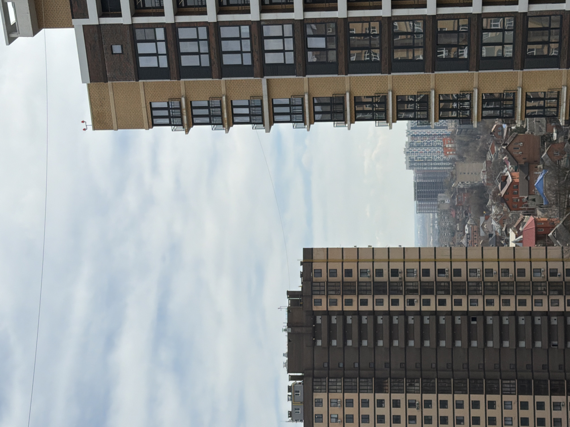 The image presents a distorted perspective of an urban landscape, likely resulting from an intentional or accidental inversion of the camera's orientation during capture.
Two multi-story buildings dominate the foreground. The upper building features a beige and brown facade with regularly spaced windows. A wire or cable stretches from the top of this building towards the sky. The lower building has a similar structure, with a beige and gray color scheme and repetitive window patterns.
Between the two buildings, a vista of an older section of the city is visible. The buildings are smaller, predominantly residential, with varying roof colors. The atmospheric conditions are slightly hazy.
The sky, exhibiting a mix of clouds and blue patches, fills the central space between the buildings. There is no visible sign of activity, people, or other moving objects within the frame.
Based on the description and known information about the city of Azov, Russia, without external information about the specific location or landmarks in the image, it is not possible to definitively identify this scene as being in Azov, Russia.