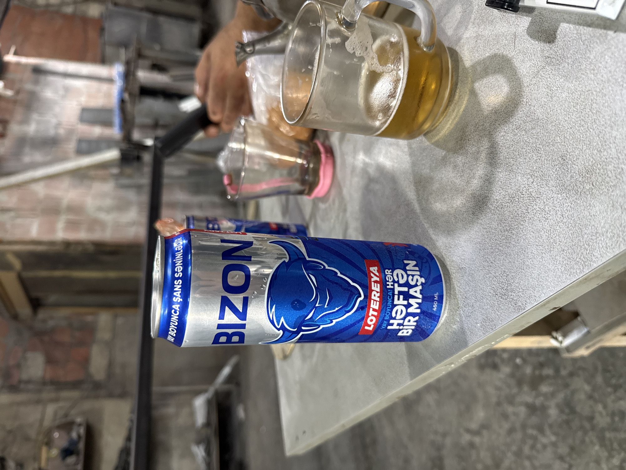 An aluminum can of "Bizon" energy drink, featuring a blue bull emblem and text in Azerbaijani indicating a lottery and weekly car prizes, sits on a textured gray surface. Adjacent to it is a glass mug half-filled with a light-colored beverage, likely beer, with a frothy head and ice cubes. In the background, out of focus, are other glasses, a plastic bag, and hands holding an object, suggesting a casual gathering. The setting appears to be an industrial or workshop environment, with visible brick walls and metal structures in the background, potentially located in Bilajari, Azerbaijan, given the text on the can.