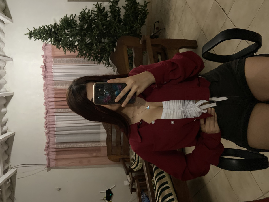 A woman sits in a black chair, taking a mirror selfie. She wears a red shirt over a white, gathered top, and dark shorts. Her phone obscures her face. The reflection shows pink-trimmed curtains, a Christmas tree, and other furniture. The floor appears to be tiled. A portion of the white, latticed ceiling is also visible in the mirror.