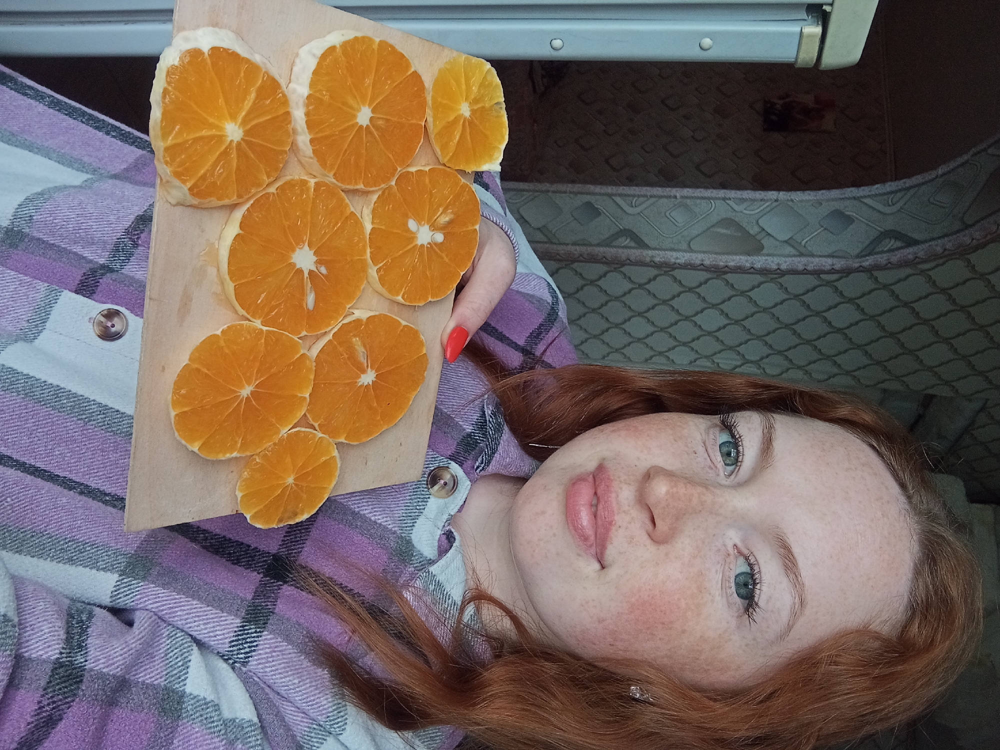 A red-haired woman with freckles is lying down indoors. She is holding a wooden board with nine sliced orange rounds on it. She is wearing a plaid shirt with white and light purple. The background includes a patterned surface and a white shelf. The location is Yurgamysh, Russia.