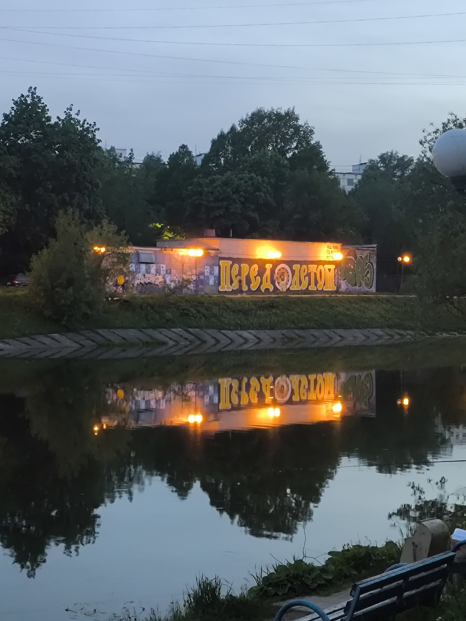 Captured at dusk, the image features a body of water reflecting an illuminated, graffiti-covered building with the word "ПЕРЕДО ЛЕТОМ" written on it in yellow. A retaining wall separates the building from the water. Trees and urban structures, including a large sphere, are visible in the background. In the foreground, a park bench sits near the water's edge. The city is unknown, Unknown.