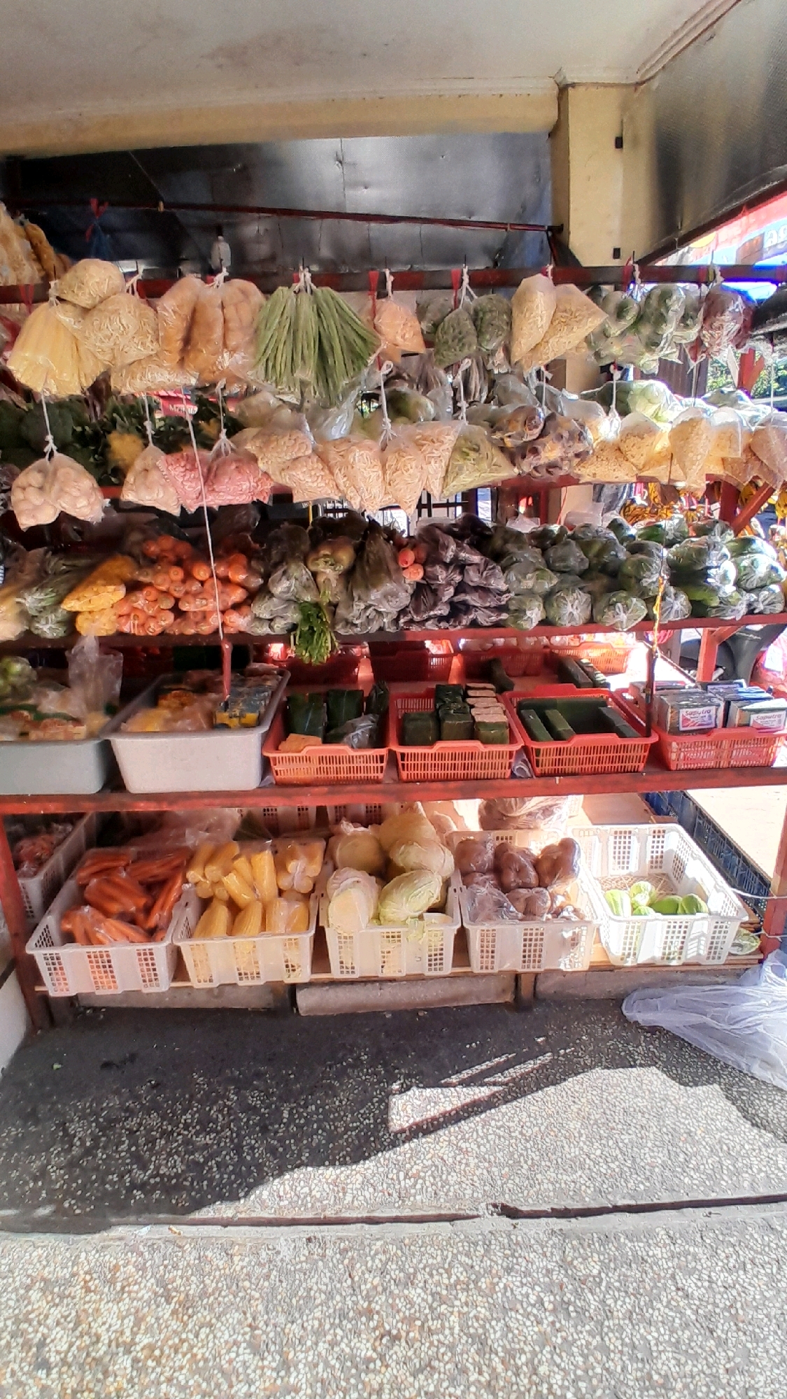 The image shows an outdoor market stall in Bandung City, Indonesia, filled with various fresh produce and packaged goods. The stall has multiple shelves stacked with items. The top shelf displays numerous plastic bags of dried goods and vegetables hanging from strings. The second shelf holds a variety of fresh vegetables and fruits, including carrots, tomatoes, corn, and leafy greens, some in plastic bags and others loose. The third shelf features containers of packaged items such as wrapped food, possibly snacks or seasonings, and more vegetables. The bottom shelf contains baskets filled with more produce, including carrots, corn on the cob, cabbages, and what appear to be onions or potatoes. The ground in front of the stall is paved with small stones and exhibits shadows from the stall structure, indicating an outdoor setting, likely on a sunny day. A white plastic bag is visible on the right side of the image on the ground. The overall scene depicts a bustling local market environment.
