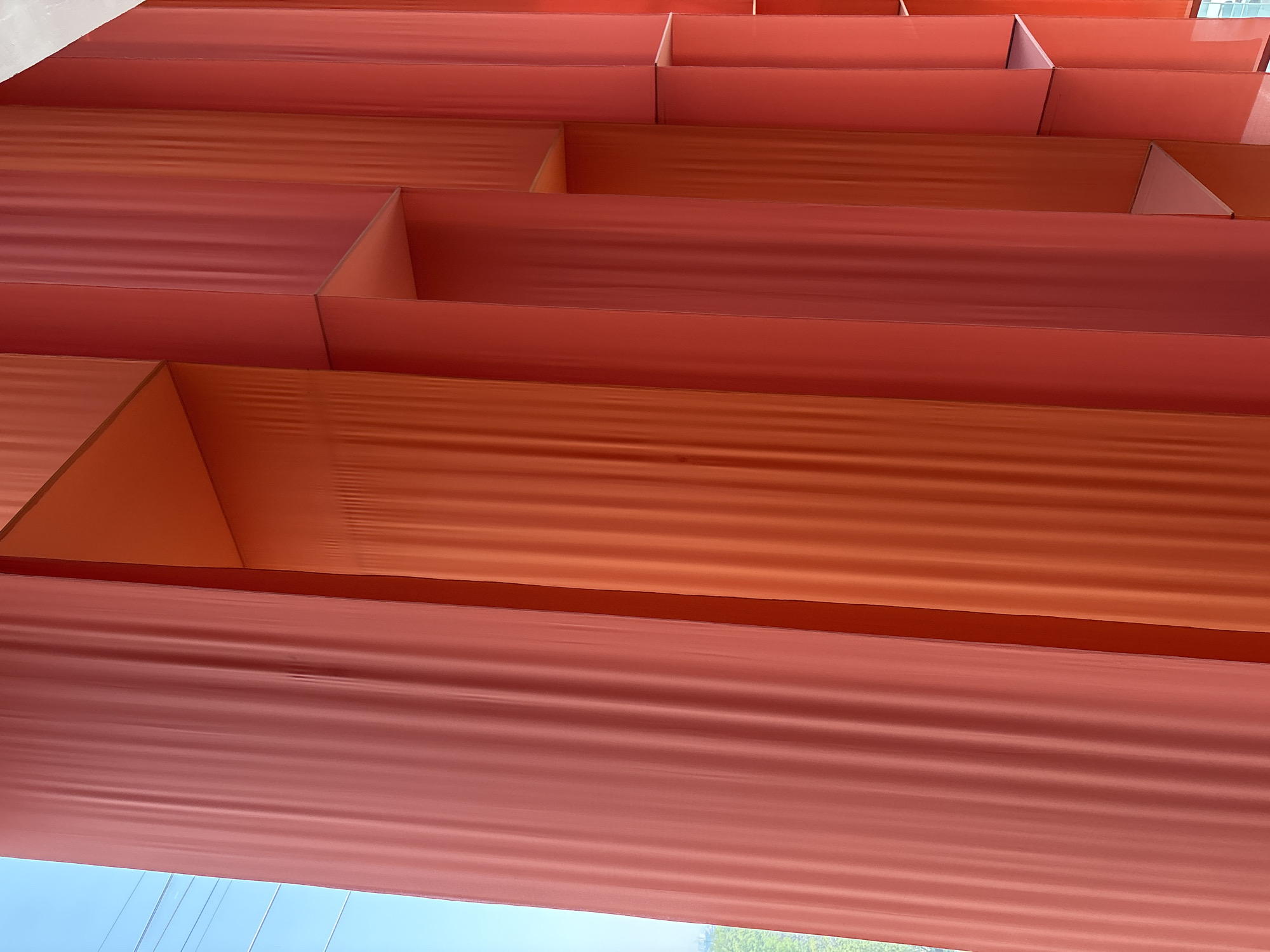 The image depicts a close-up, low-angle view of a series of architectural structures, possibly the facade or detail of a building in Mexico City, Mexico. The structures appear to be a series of rectangular boxes or containers, stacked in a stepped or layered fashion. They are constructed from a material that is uniformly colored in various shades of orange and red, with a visible corrugated or ridged texture, particularly on the flat surfaces.
The perspective is angled upwards, revealing a glimpse of the outside environment, including a portion of the sky which transitions from a lighter, bluish-white color towards the bottom left corner, suggesting natural daylight. Hints of greenery are also visible near the sky.
The lighting suggests an outdoor environment with indirect sunlight, emphasizing the texture of the materials and creating shadows within the recessed areas of the boxes. The entire scene presents an interplay of architectural design, texture, color, and light.