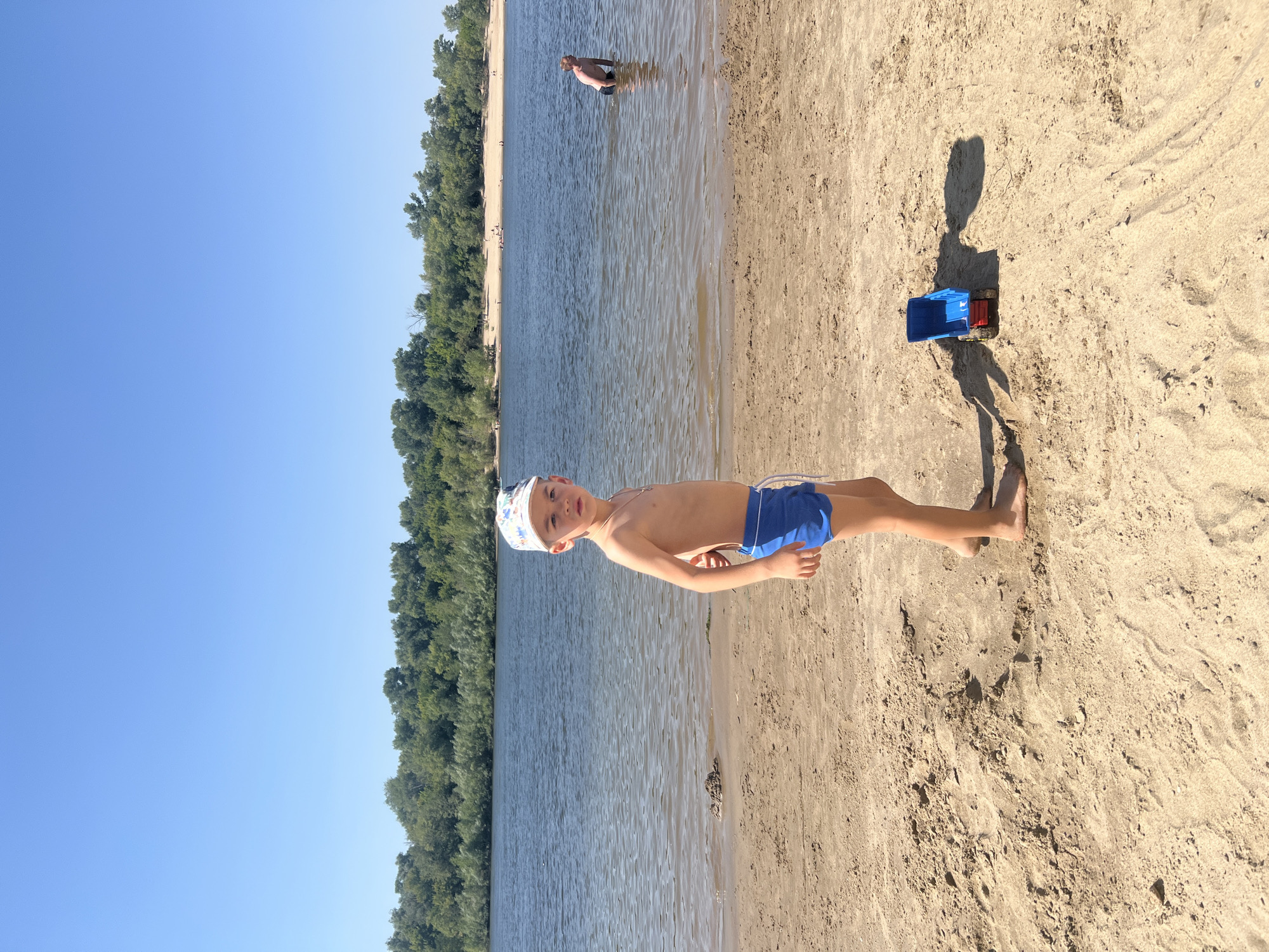 A young boy wearing blue swim trunks and a white patterned baseball cap stands on a sandy beach, looking towards the camera. He is positioned slightly off-center to the right. To his right, a small blue toy truck is partially visible in the sand. The beach is wet near the water's edge. In the background, a wide river stretches across the frame, with a densely wooded bank on the far side. The sky is clear and blue. Another person is visible in the water in the far background, to the right. The overall scene depicts a sunny day at a river beach. The location is stated to be the city of Kilyakovka, Russia.