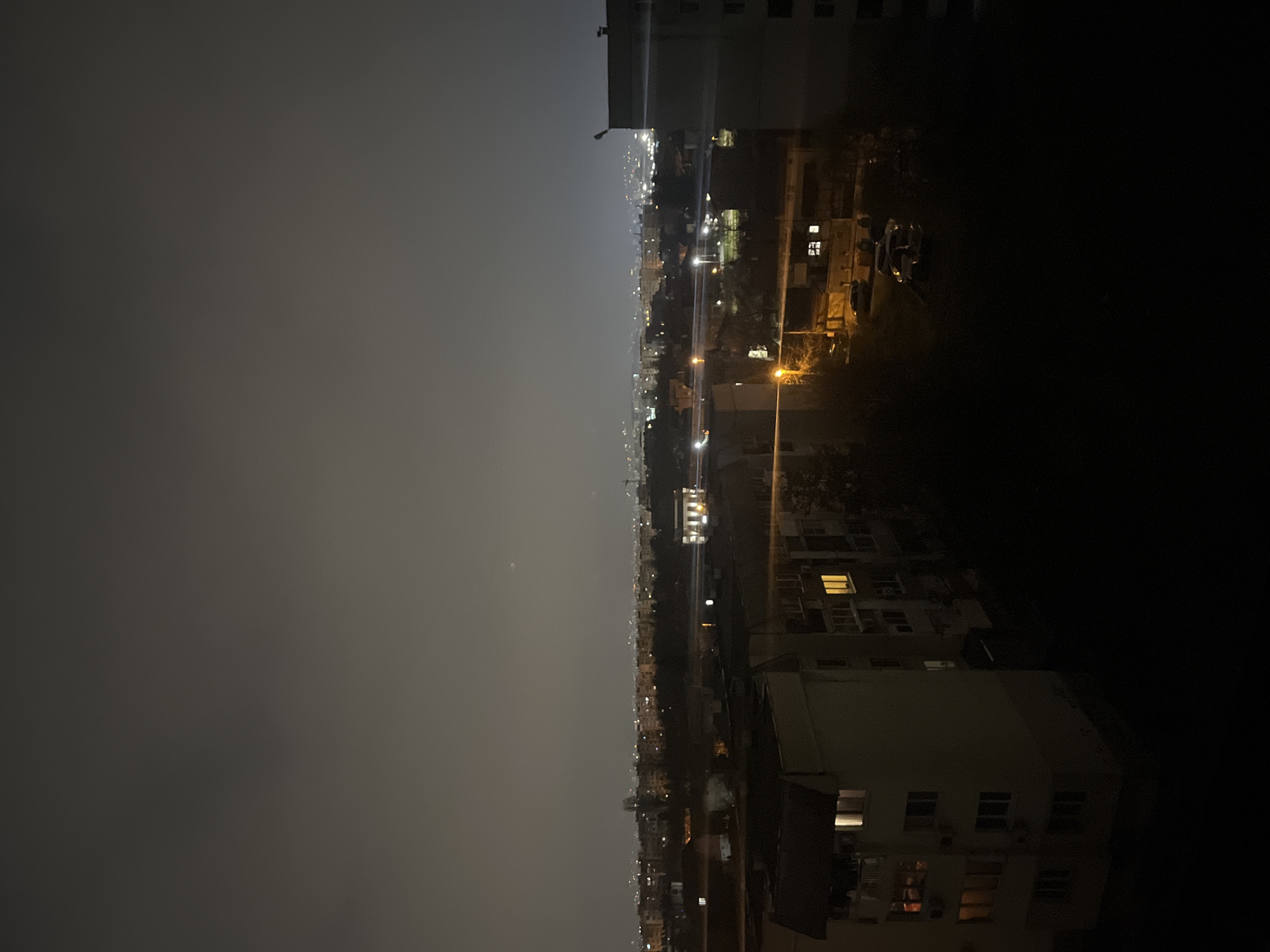 The image depicts a nighttime view of Sofia, Bulgaria, from an elevated position, likely a high-rise building. The scene shows a cityscape extending into the distance under a dark, overcast sky. City lights illuminate the streets and buildings. Closer to the foreground, there are buildings with illuminated windows. The right side of the image is darker, with the silhouette of a nearby building obscuring details. The overall atmosphere is that of a quiet, urban night scene.