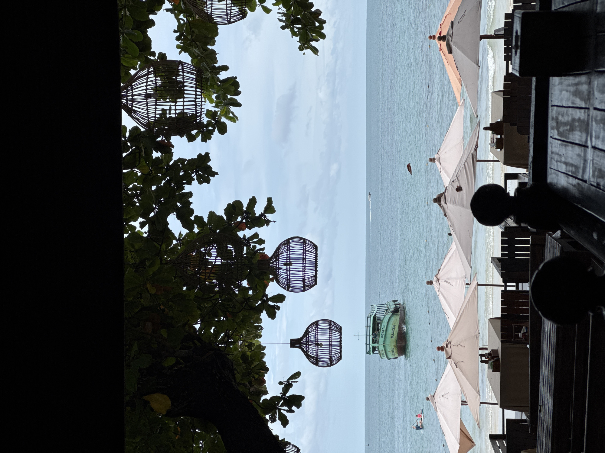 The image captures a view from an outdoor seating area overlooking Ao Phai beach in Ko Samet, Thailand. In the foreground, dark wooden tables and chairs are arranged, some with dark, silhouetted figures. Behind the seating area, several beach umbrellas are set up, with tables underneath them. The umbrellas are a mix of beige and a light orange color.
Further back, the turquoise ocean stretches out under a cloudy sky. A green boat with the name "NARUMOL" on its side is visible on the water, heading towards the viewer. Two people are on a paddleboard in the distance, to the left of the boat. Overhead, the branches of a tree frame the scene, with several decorative woven lanterns hanging from them. The overall impression is a relaxed, beachfront setting.