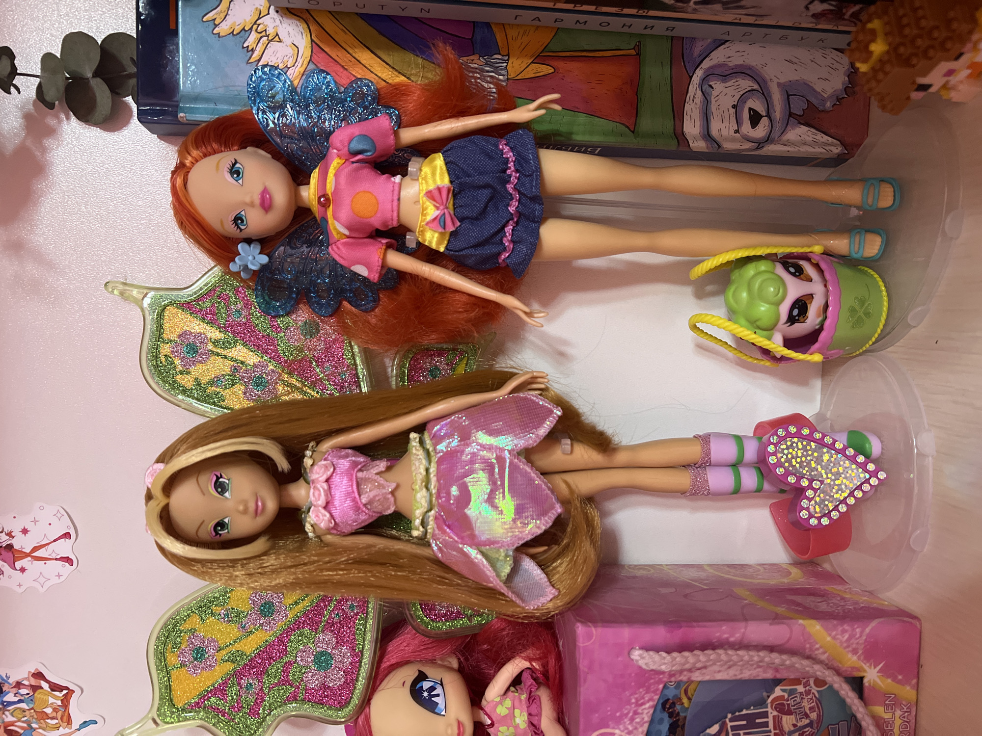 This is a close-up, eye-level shot of two Winx Club dolls standing on a clear plastic display base. The doll on the left has long, blonde hair with bangs and wears a pink floral top and a iridescent pink skirt with green trim. She has large, glittery green wings. The doll on the right has long, red hair and wears a pink, yellow, and blue outfit with blue wings. She is wearing blue sandals.
To the left of the dolls is a pink box with a rope handle and a picture on the front. A small doll with pink hair is partially visible behind the pink box. In front of the dolls, there is a pink heart-shaped accessory with rhinestones and a small green basket with a sheep toy. Behind the dolls are some stacked books or boxes, one of which has an illustration of a polar bear. The background is a pink wall with some cartoon stickers.
This image does not contain any information that can be used to determine its location within the city of Shymkent, Kazakhstan.