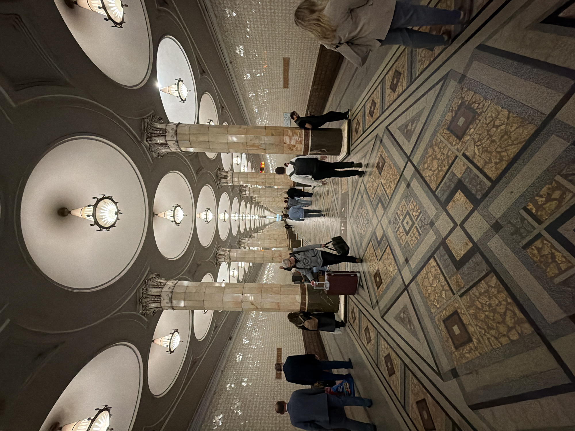 The image depicts a platform in a Moscow, Russia metro station. The platform features a patterned floor with geometric designs. Rows of ornate columns extend into the distance, supporting a ceiling with evenly spaced, circular light fixtures. Several people are standing and walking on the platform, with one person leaning against a column. Some individuals are pulling luggage. The overall lighting is dim, enhancing the architectural details of the station.