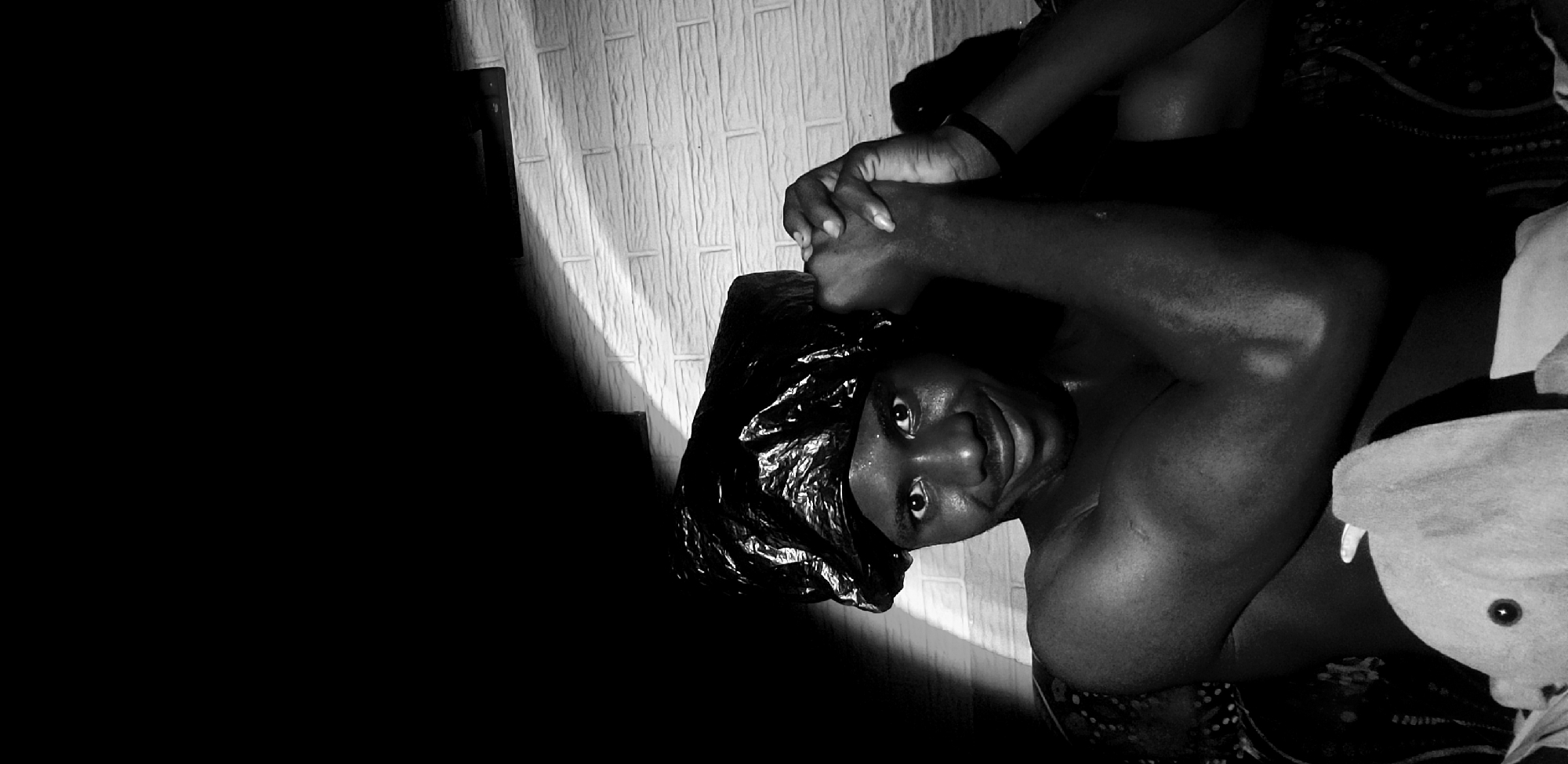 A black and white, medium close-up shot shows a young Black man with a plastic bag wrapped around his head, his hands clasped in front of his face. The man is shirtless, with visible sweat on his skin, suggesting a warm environment. He has dark eyes and a slight smile. The background is dimly lit, with a textured wall that appears to be made of white bricks. A shadow falls across the wall from the upper left of the frame, creating a stark contrast and highlighting the subject. The overall mood is intimate and perhaps playful. There is no specific indication of the city of Narayi, Nigeria.