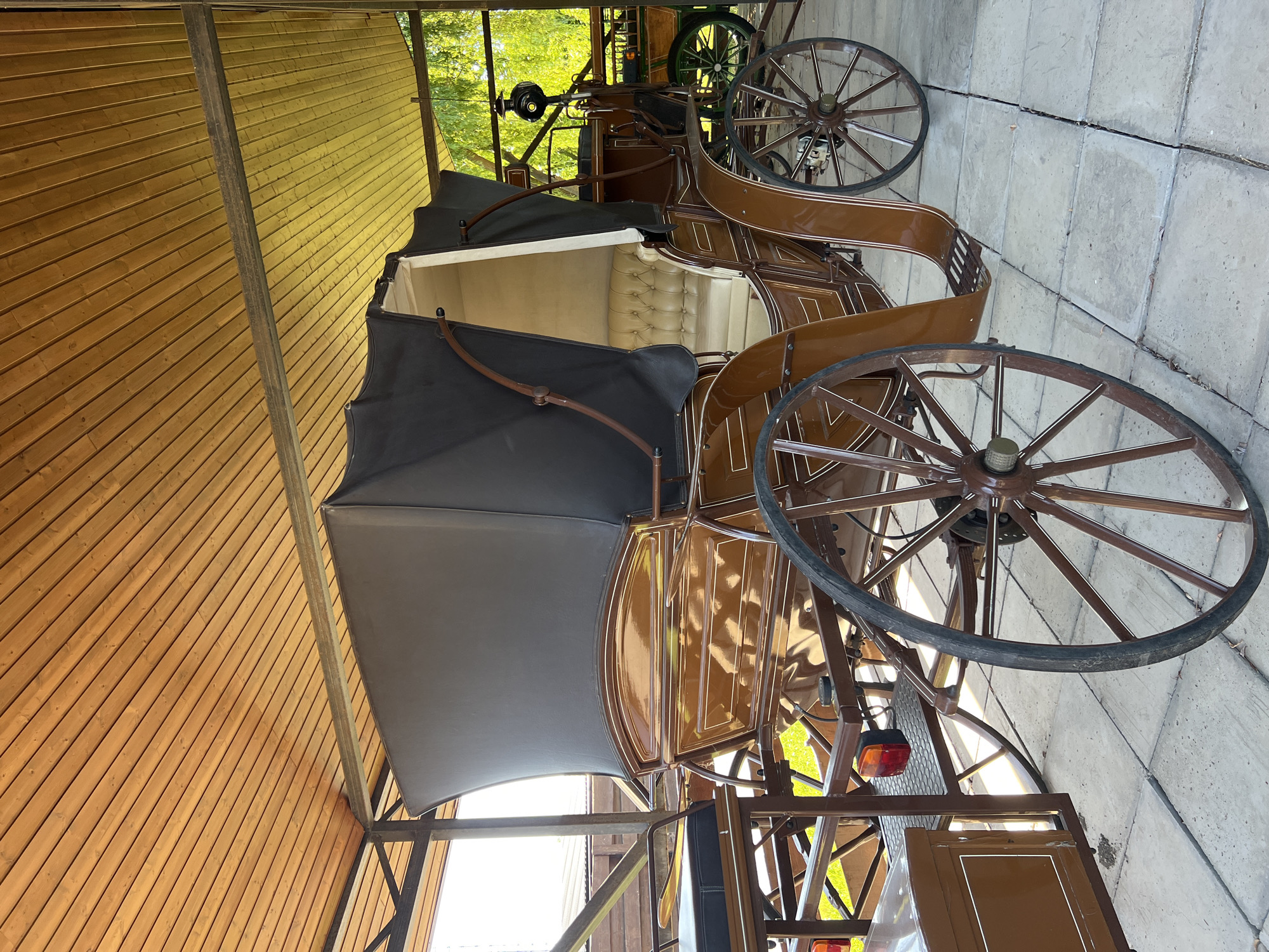 The image shows a brown horse-drawn carriage with a black canopy parked under a wooden-slatted roof. The carriage has two large spoked wheels on each side and a padded interior with light-colored upholstery. There is a second, partially visible carriage in the background, which appears to be green and has a similar wheel design. The carriages are parked on a paved surface made of light-colored square tiles. The scene appears to be outdoors, with greenery and trees visible behind the carriages, suggesting a park or historical site. The question asks for the location in Plastunivska, Russia, but no visual elements in the image confirm this specific location.