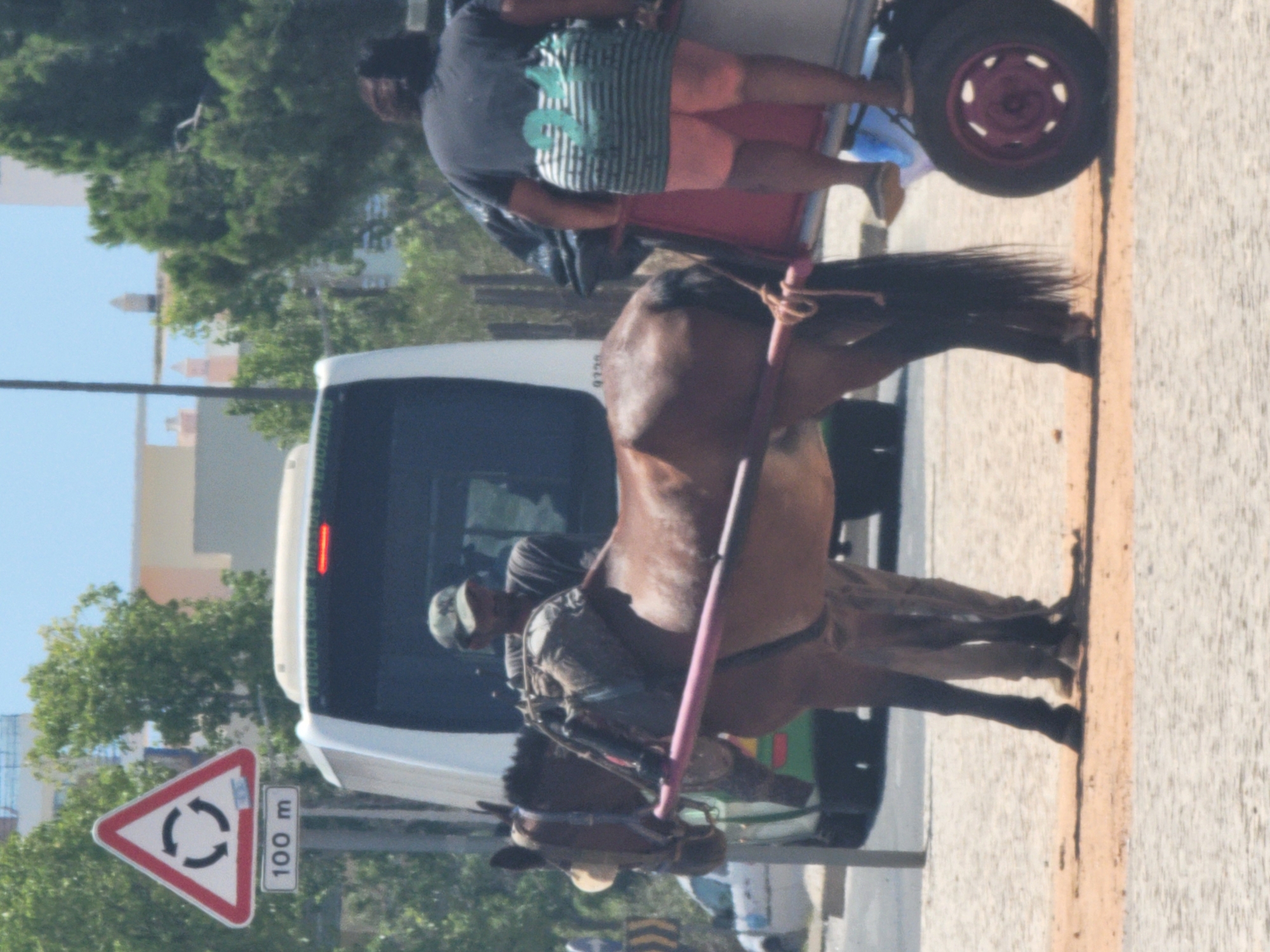 In Quarteira, Portugal, on a sunlit day, a brown horse is harnessed to a red cart. The horse, wearing a blue halter and a saddlebag filled with items, stands on a sandy surface. A man wearing a camouflage baseball cap and a dark t-shirt, likely the driver, stands beside the horse. Behind them, a white bus is parked. Another person, seen from the back, is sitting on the red cart, wearing a dark t-shirt with a green graphic and striped shorts. A traffic sign with a circular arrow symbol and "100 m" below it is visible to the left of the scene, indicating a roundabout ahead. Trees and buildings are visible in the background.