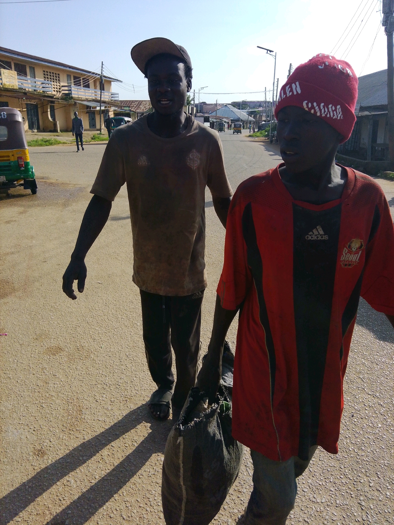 The image captures a street scene in Jos, Nigeria, under a bright, clear sky. In the foreground, two young Black males walk towards the camera. The male on the left wears a brown t-shirt and black pants, with a brown baseball cap tilted back. He has his left arm extended, hand open. The male on the right wears a red and black striped t-shirt and grey pants. He has a red beanie on his head and is carrying a large, dark-colored garbage bag in his right hand. The ground is an asphalt road with a rough texture.
In the background, a light-colored, two-story building with balconies and numerous windows lines the street on the left. A small, yellow and green motorized tricycle, commonly known as a Keke Napep, is parked on the side of the road near the building. Further down the street, other buildings and structures are visible, along with utility poles and electrical wires crisscrossing the sky. The overall atmosphere suggests a casual daytime activity on a city street.