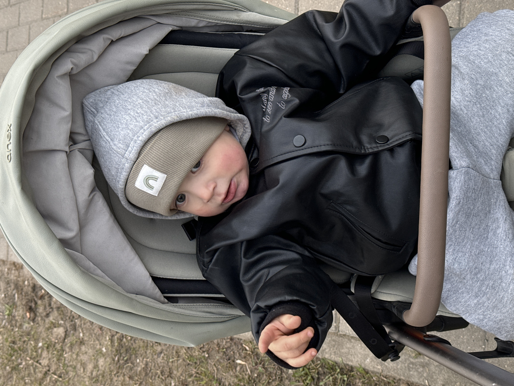A baby or young toddler is pictured lying in a light olive-green stroller, looking up towards the camera with a curious and slightly smiling expression. The child is dressed in a light grey hooded top, a beige knitted beanie adorned with a small white square patch featuring a pastel rainbow logo, a black faux leather jacket, and light grey sweatpants. The baby's cheeks appear rosy.
The stroller has a padded grey interior, and a brown-colored handlebar is visible on the right side of the frame. The background and foreground suggest an outdoor setting, possibly on a paved path or sidewalk, with patches of dirt or dry grass visible. The lighting indicates it is daytime.
Visible text includes a stylized, vertically embossed "XOUX" logo on the side of the stroller. On the right shoulder of the black jacket, white text is partially visible, reading "many to see and to expl," suggesting a phrase like "many things to see and to explore." The location context is Akulovo (Gp Kubinka), Russia.