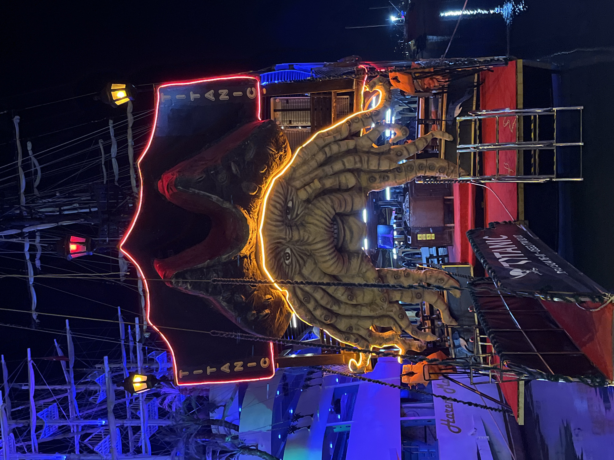 This nighttime image features a large, elaborately decorated ship, likely a themed attraction or party boat, in Alanya, Turkey. The primary subject is the ship itself, distinguished by a massive, sculpted kraken or octopus figure prominently attached to its side. This kraken sculpture is dramatically outlined with bright orange neon lights, highlighting its tentacles and a large eye, making it glow against the dark background.
The ship is extensively illuminated with artificial lights. Red neon lights trace the outline of the ship's hull and upper structures, while blue lights cast a glow on the masts, rigging, and some deck areas. Several yellow-lit lanterns hang from the ship's masts, contributing to the maritime aesthetic.
Visible text on the ship includes "TITANIC" in large, illuminated red letters, appearing twice on different sections. A banner hanging from the ship also displays "TITANIC" along with the text "DC 0304" and a small anchor-like symbol. Partially visible text "Harem M" can be seen on a white section of the boat.
The scene is set at night, with the dark sky and water emphasizing the bright, colorful illumination of the ship. Reflections of the lights are visible on the water in the background, indicating the vessel is either docked or cruising. The overall impression is that of a fantastical, visually striking nautical spectacle.