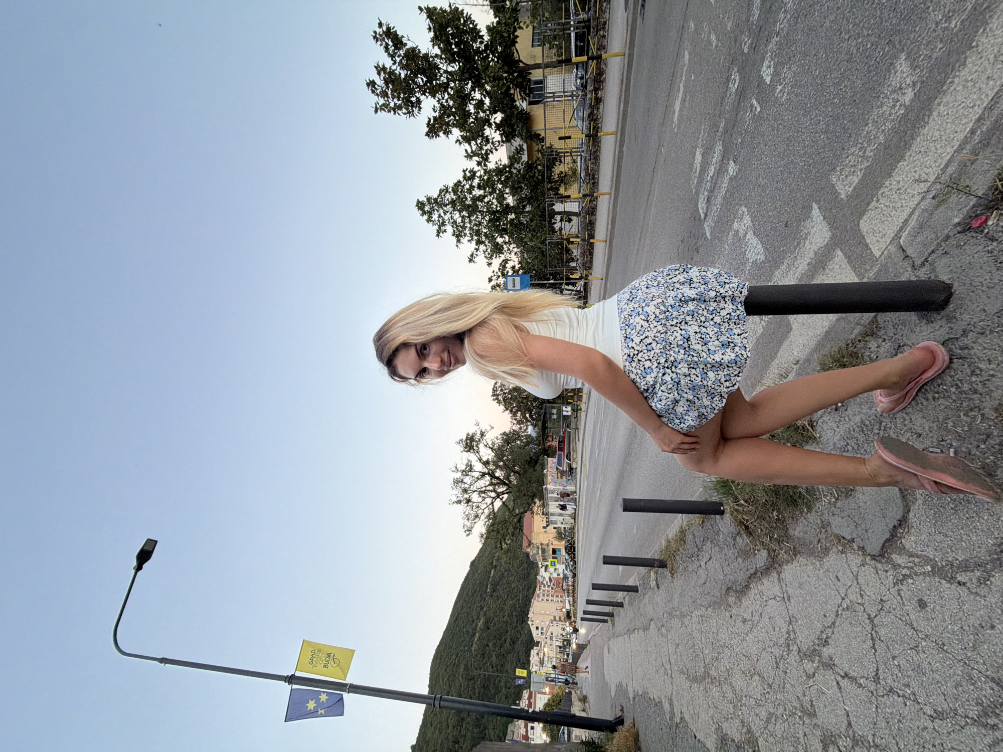 A woman with blonde hair is sitting on a black pole next to a road in Budva, Montenegro. She is wearing a white tank top and a floral skirt. The road has a crosswalk on it, and there are cars and buildings in the background. The sky is clear and blue. There are two flags on a lamppost to the left of the woman. One flag is blue with yellow stars, and the other is yellow with writing on it. The overall scene suggests a casual, outdoor setting in a European city.