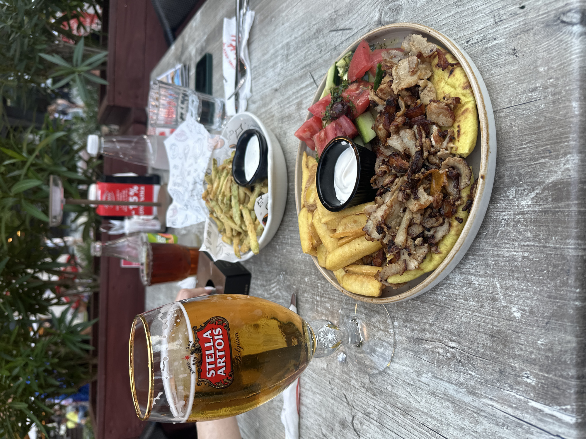 A table at an outdoor restaurant in Varna, Bulgaria, displays a meal. In the foreground, a glass of Stella Artois beer and a plate of gyro meat with fries, salad, and a dollop of white sauce are arranged on a wooden table. A white bowl filled with fried green beans and a small dish of white sauce is visible in the background, along with a bottle of water and a red carton. Lush green plants are in the background, suggesting an outdoor setting. The lighting indicates it is daytime.