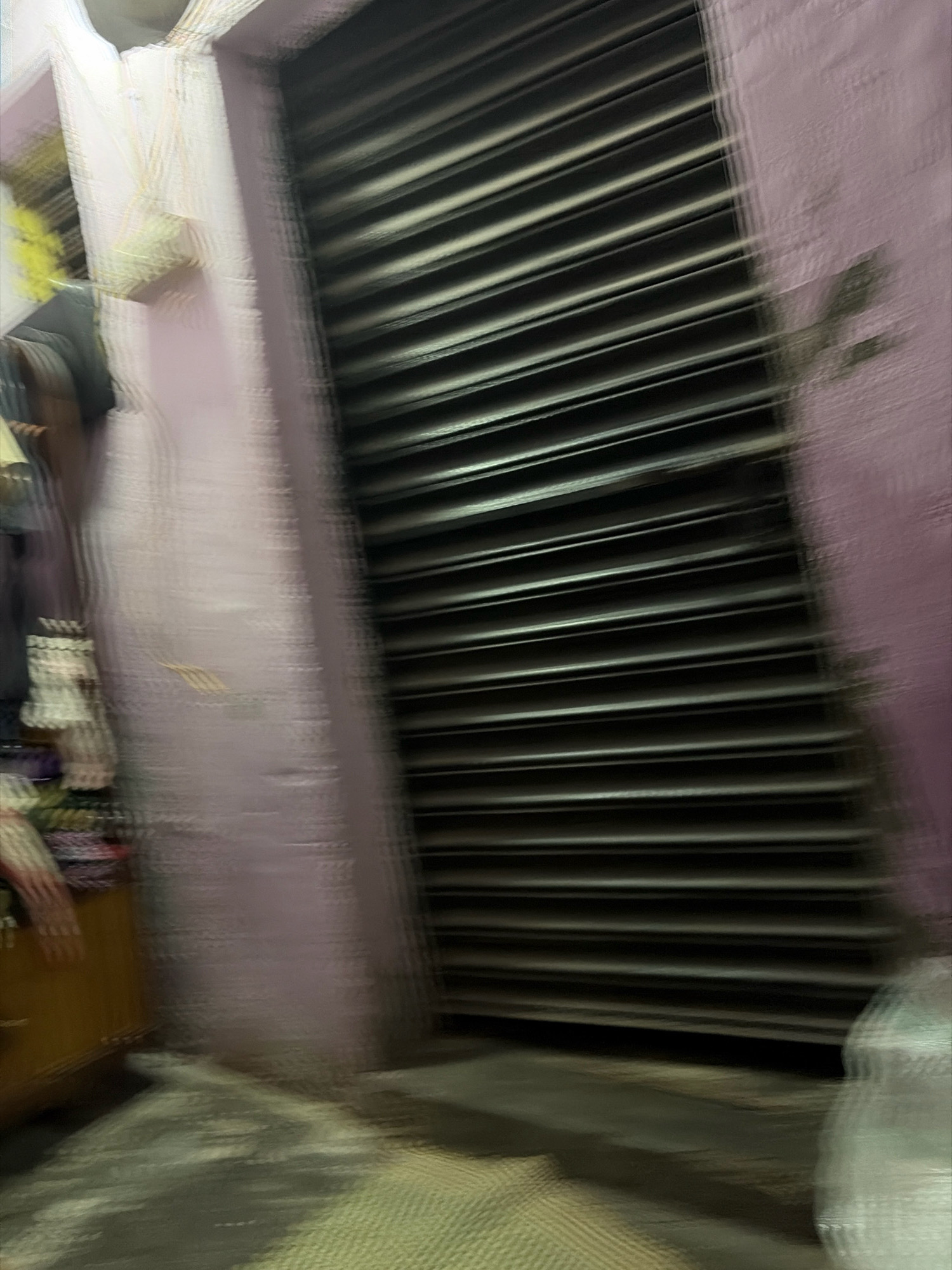 The image depicts a dimly lit, urban scene, likely in Jamshedpur, India, characterized by a blurred motion effect that suggests movement or a quick capture. In the foreground, a person's hand and arm are visible, resting on a wooden surface that appears to be a part of a small table or cart. The person is wearing striped clothing on their arm. Behind the hand, a section of a light pink wall is visible, with some indistinct hanging items and a yellow object. To the right of the wall is a dark, ribbed shutter, possibly of a shop or building entrance, which dominates a significant portion of the frame. The ground in front of the shutter and wall appears to be paved with textured tiles or concrete. A blurred white object, possibly a bag or piece of fabric, is present in the lower right corner. The overall atmosphere is somewhat hazy and indistinct due to the motion blur.