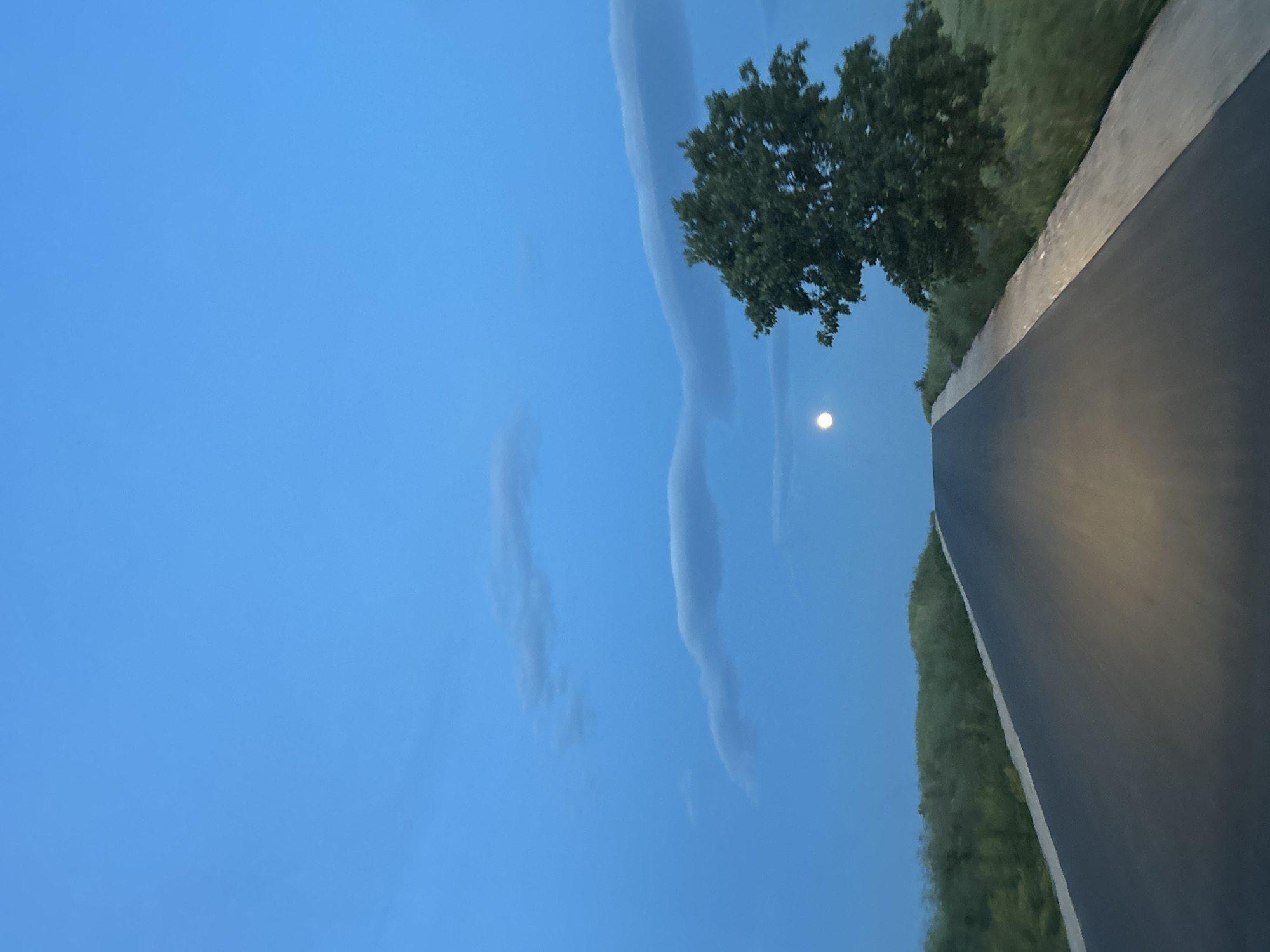 The image depicts a road scene at twilight in Smilets, Bulgaria. The sky is a light blue with wispy clouds. A full moon is visible near a tree with green foliage. Adjacent to the tree is a paved road with grassy verges on both sides. The road surface is dark and reflects light. The image is rotated.