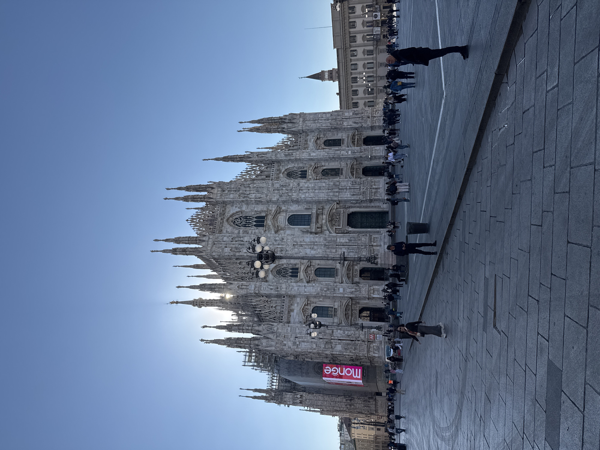 The image captures Milan Cathedral, Italy, from ground level. A large group of people are present near the cathedral. To the side of the cathedral is a paved square, with people walking and standing around. The sky above is clear and blue. An advertising sign is visible on one side of the cathedral.