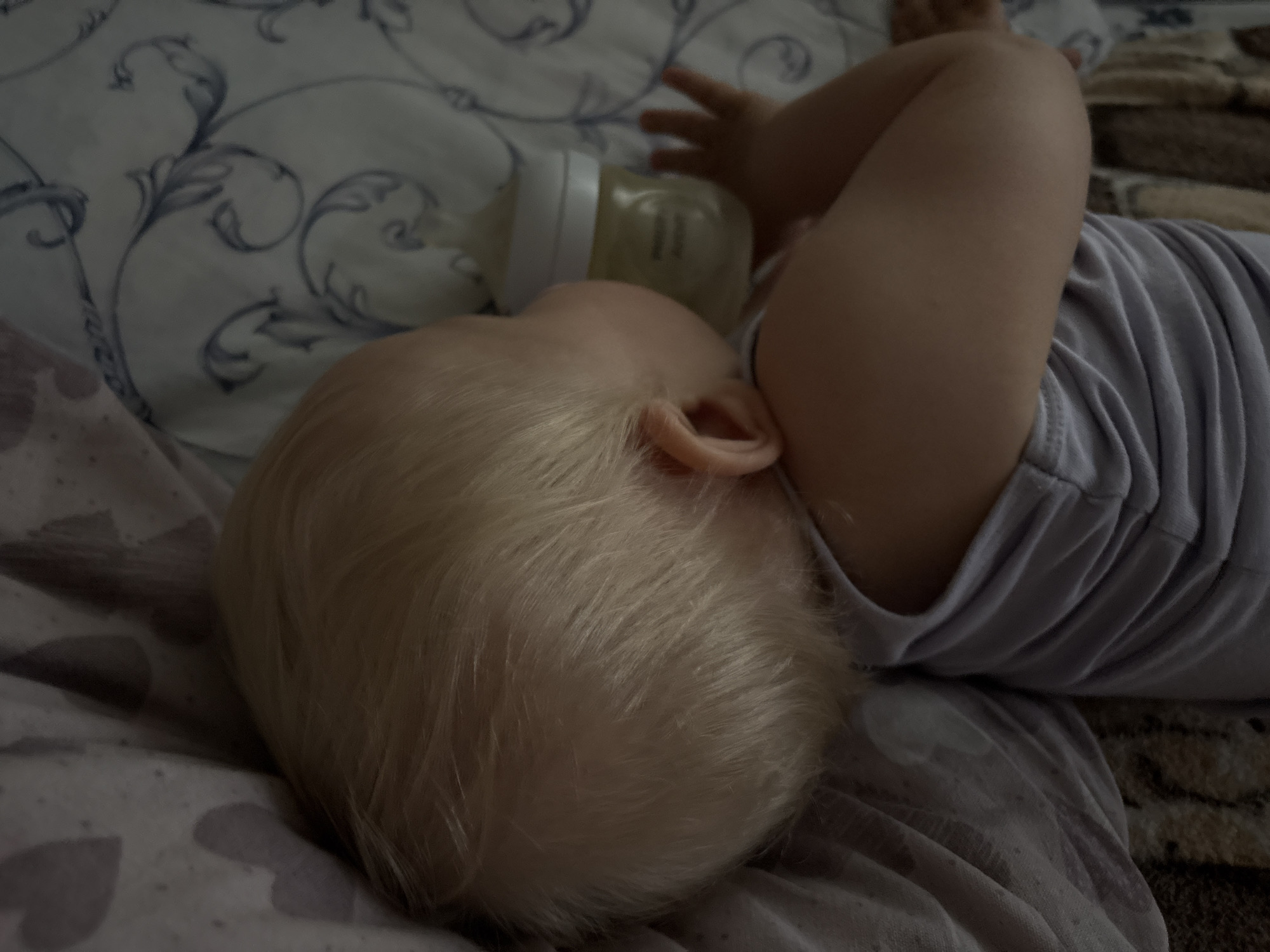 A baby with blond hair is sleeping on a bed. The baby is lying on their side, facing away from the camera, with a baby bottle next to their head. The baby is wearing a light purple t-shirt. The bed is covered with a patterned sheet and a textured blanket. The scene appears to be indoors, likely in a bedroom, and there is no information that would place it in Sovkhoza "Razdol'e", Russia.