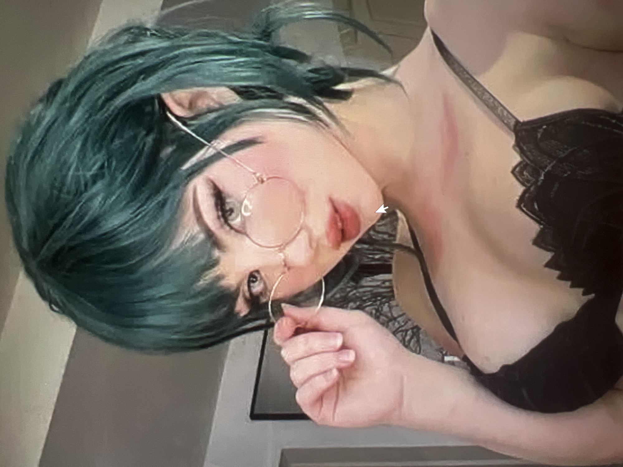 The image shows a close-up, waist-up shot of a person with teal-colored hair, wearing large, round, rose-tinted glasses and a black lace bra. The person's left hand is raised, holding the rim of the glasses. The background is indistinct and appears to be an indoor setting with a wall and a framed picture visible. There are no discernible elements in the image that would allow for the identification of the location as Minsk, Belarus.