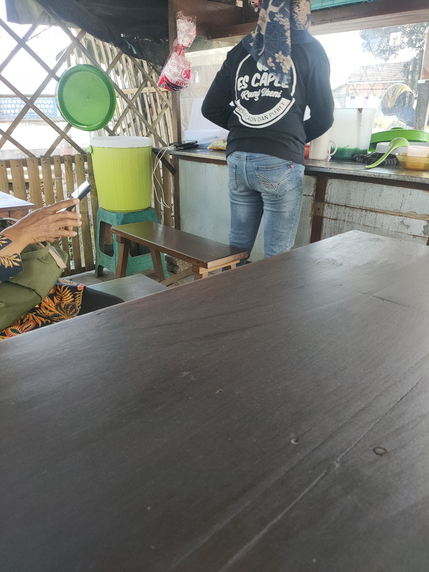 The image depicts an outdoor scene at what appears to be a food stall in Kanigoro, Indonesia. In the foreground, a dark wooden table occupies much of the frame, showing signs of wear. To the left, a person's hands are visible, holding a smartphone. The person is wearing a green and patterned garment. Behind them, a bright green cooler with a matching lid sits on a plastic stool. Further back, a person wearing a black long-sleeved shirt with white text and blue jeans stands behind a counter. The shirt has a circular logo with text that reads "ES CAPLE RANI DHERI PESICAON DAN PLERET". The counter is cluttered with various items, including cups, a pitcher, a green ladle, and a bowl. The background shows a fence made of bamboo or wood, and beyond that, a building and trees are visible under a bright sky. The overall impression is of a casual, open-air establishment.