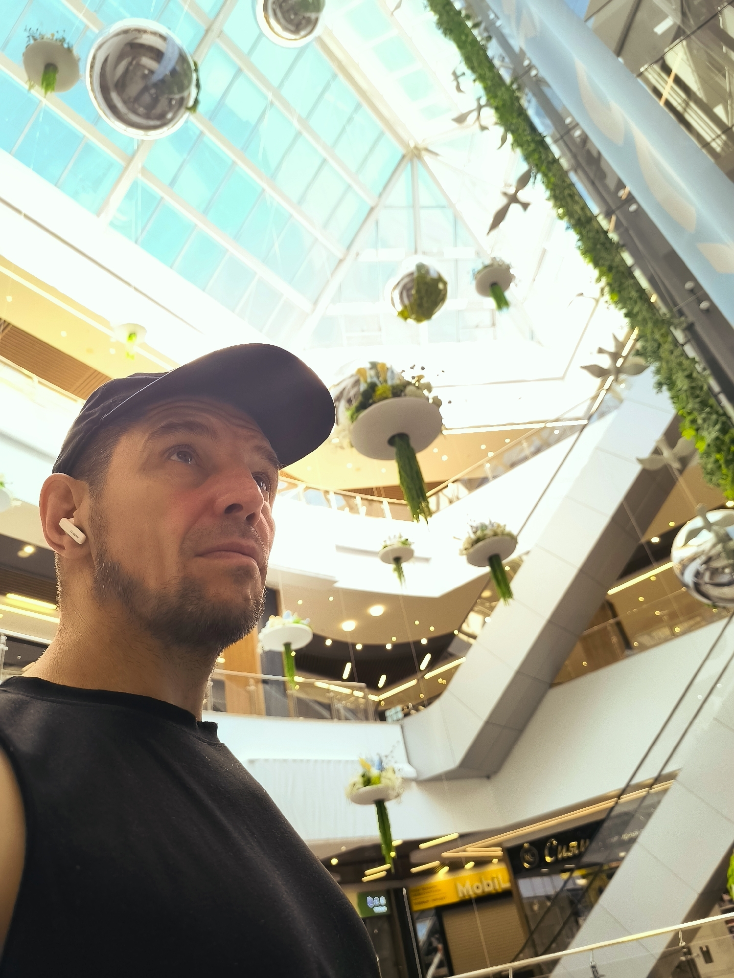 The image shows a man wearing a black t-shirt and a black baseball cap, looking upwards and to the right. He has a short beard and is wearing a white wireless earbud in his right ear. The background is a bright, modern interior with a large, angular atrium. Escalators and walkways are visible on multiple levels. Decorative elements, including suspended circular planters with greenery and silver spheres, are hanging from the ceiling. The architecture features a glass ceiling allowing natural light to enter. The scene appears to be inside a shopping mall or a similar large public building. The text "Mobil" is visible on a yellow sign in the background. This image was taken in Qarağandı city administration, Kazakhstan.
