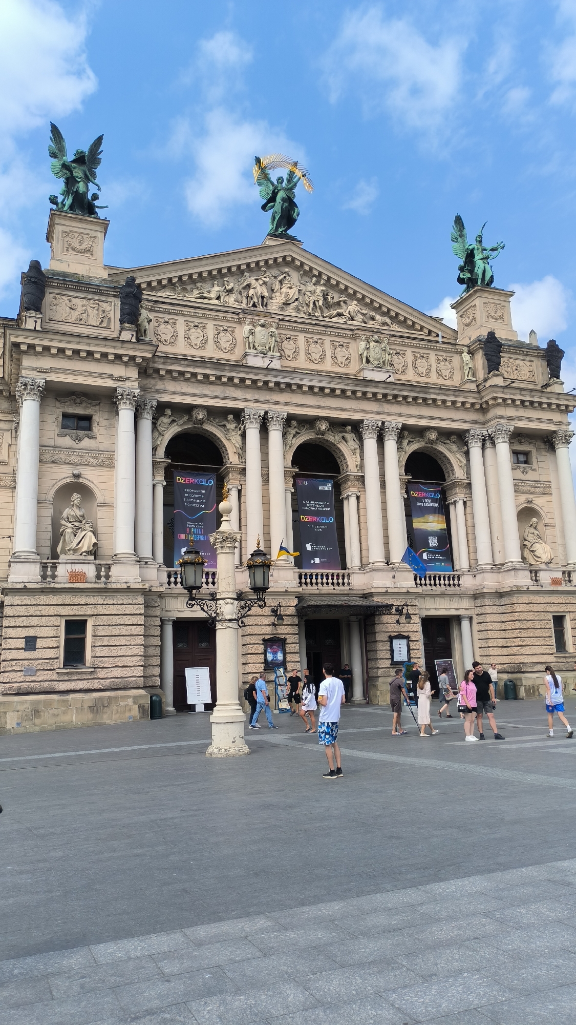 The image shows the Lviv Opera House in Lviv, Ukraine. The grand building features ornate architecture with statues and columns. Two large banners hang from the facade, displaying text and graphics. A street lamp is positioned in the foreground, and several people are walking in the square in front of the building. The sky above is blue with scattered clouds.