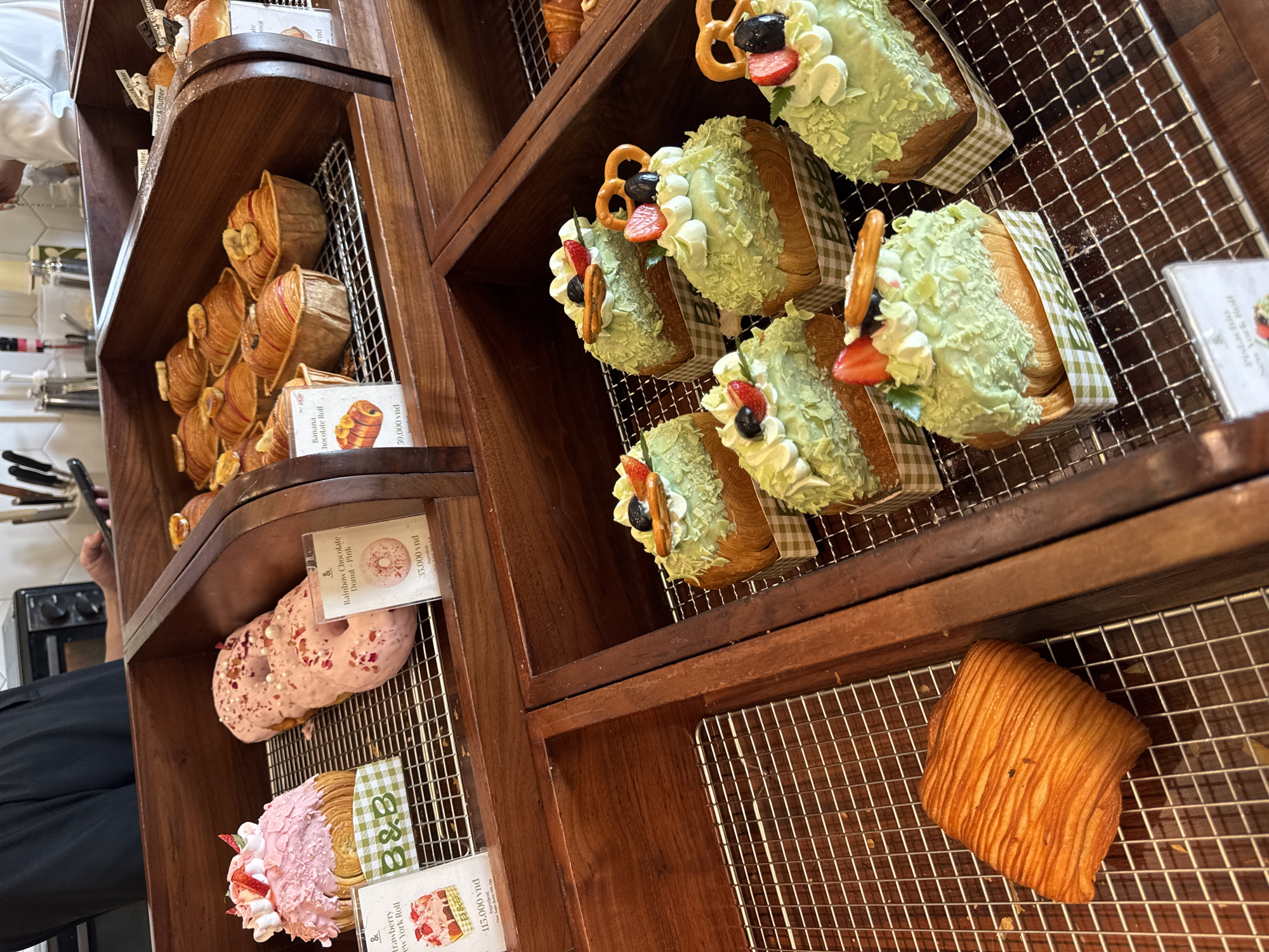 The image depicts a display case in a bakery, likely in Đà Nẵng, Vietnam, given the currency displayed on the price tags. The wooden shelves hold various pastries arranged on wire racks. The top shelf features pastries topped with green frosting, pretzels, strawberries, and blueberries, wrapped in a checkered paper. The middle shelf holds croissants and pink donuts. The bottom shelf displays a strawberry topped pastry and a single, unadorned croissant. Price tags are visible with VND (Vietnamese Dong) currency amounts. A person in a white uniform is partially visible on the left side of the frame, and kitchen equipment can be seen in the background.
