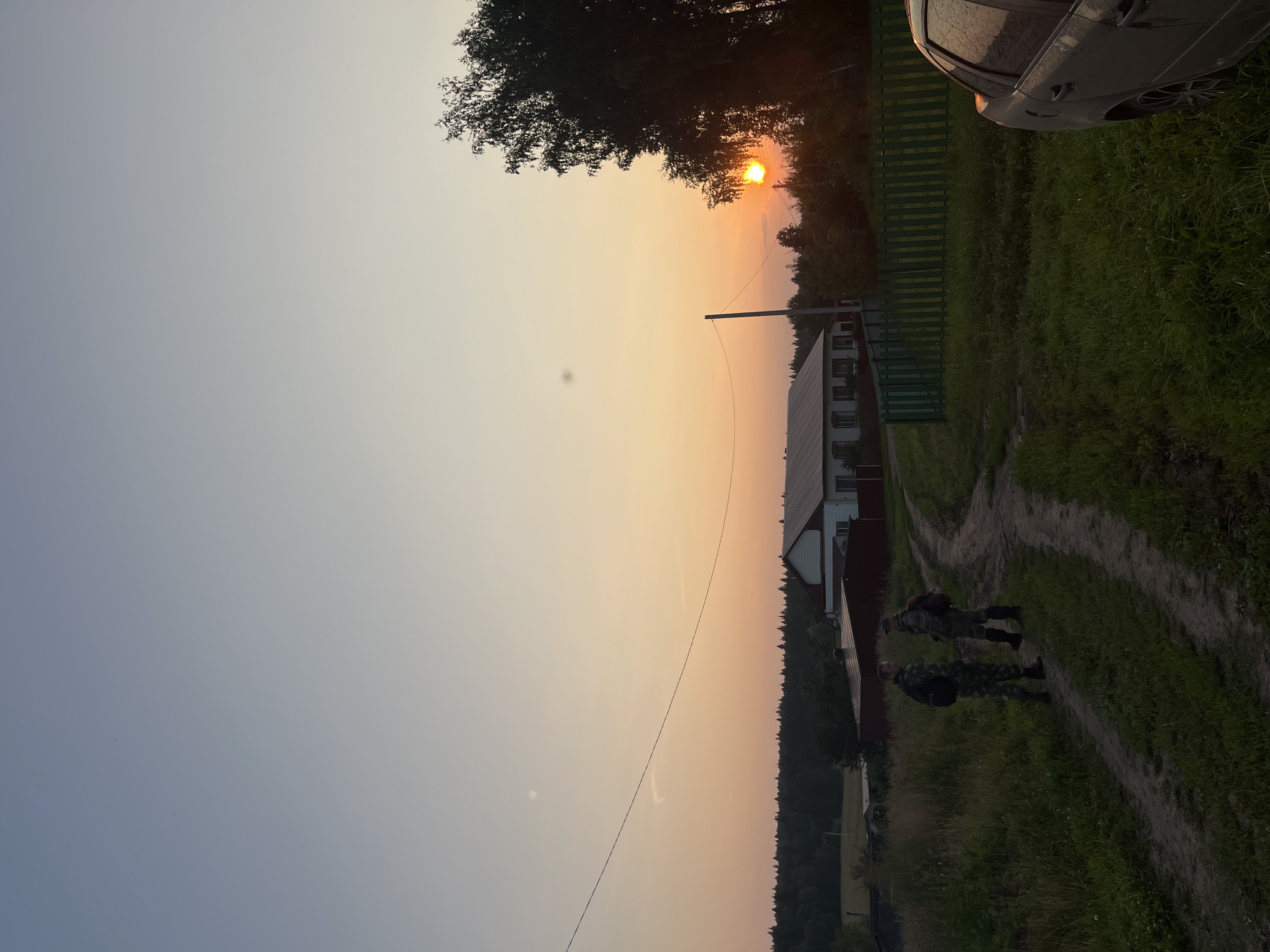 The image depicts a rural scene at sunset in the village of Perebor, Russia. Two individuals in camouflage uniforms are standing on a dirt path in a grassy area. Behind them, a white house with a red roof is visible, bordered by a green wooden fence. To the right of the scene, the sun is setting behind trees, casting a warm glow. A portion of a dark-colored car is parked on the right side of the frame, with its rear end facing the viewer. The sky exhibits a gradient of colors, transitioning from a pale blue at the top to orange and pink hues near the horizon. There are utility poles and wires present in the middle ground. The overall environment suggests a peaceful, rural setting during dusk.