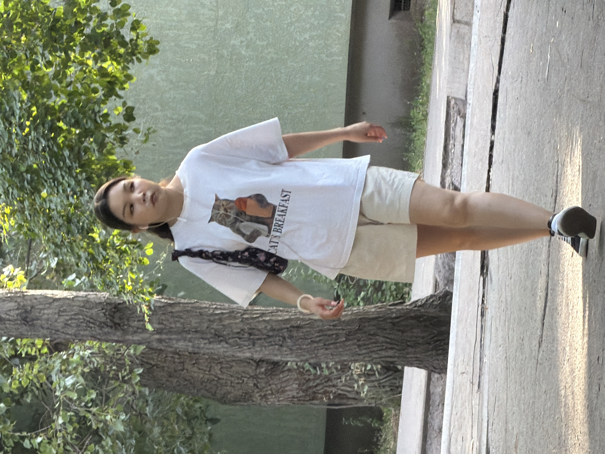 An Asian female walks outdoors in a city. She has brown hair pulled back and is wearing a white t-shirt with a graphic of a cat holding a milk carton, light-colored shorts, black and white sneakers, and white earbuds. She is carrying a small dark purse with a floral pattern over her left shoulder. A tree trunk is to her left, and she is walking on a paved surface. The background consists of green foliage and a light green building. The time of day appears to be daytime with sunlight casting shadows.