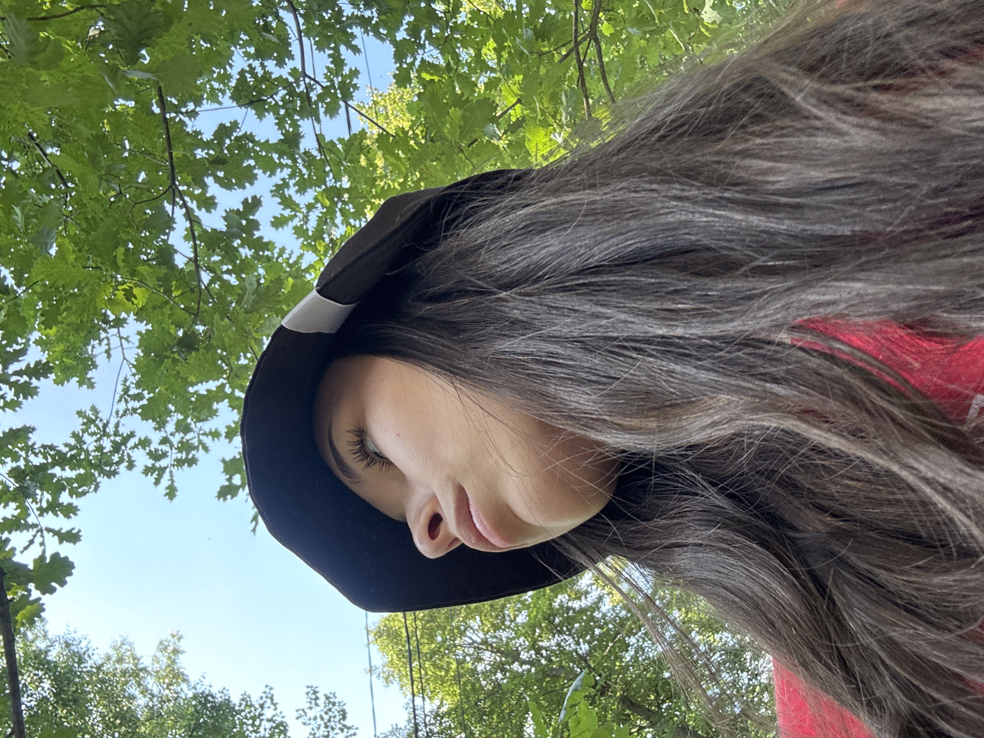 A low-angle shot shows a young woman wearing a black bucket hat and a red top, looking upwards. She has long, dark brown hair and her face is partially obscured by the hat. The background consists of green foliage and a blue sky, suggesting an outdoor setting. The image was taken in Doma 1096 Km, Russia.