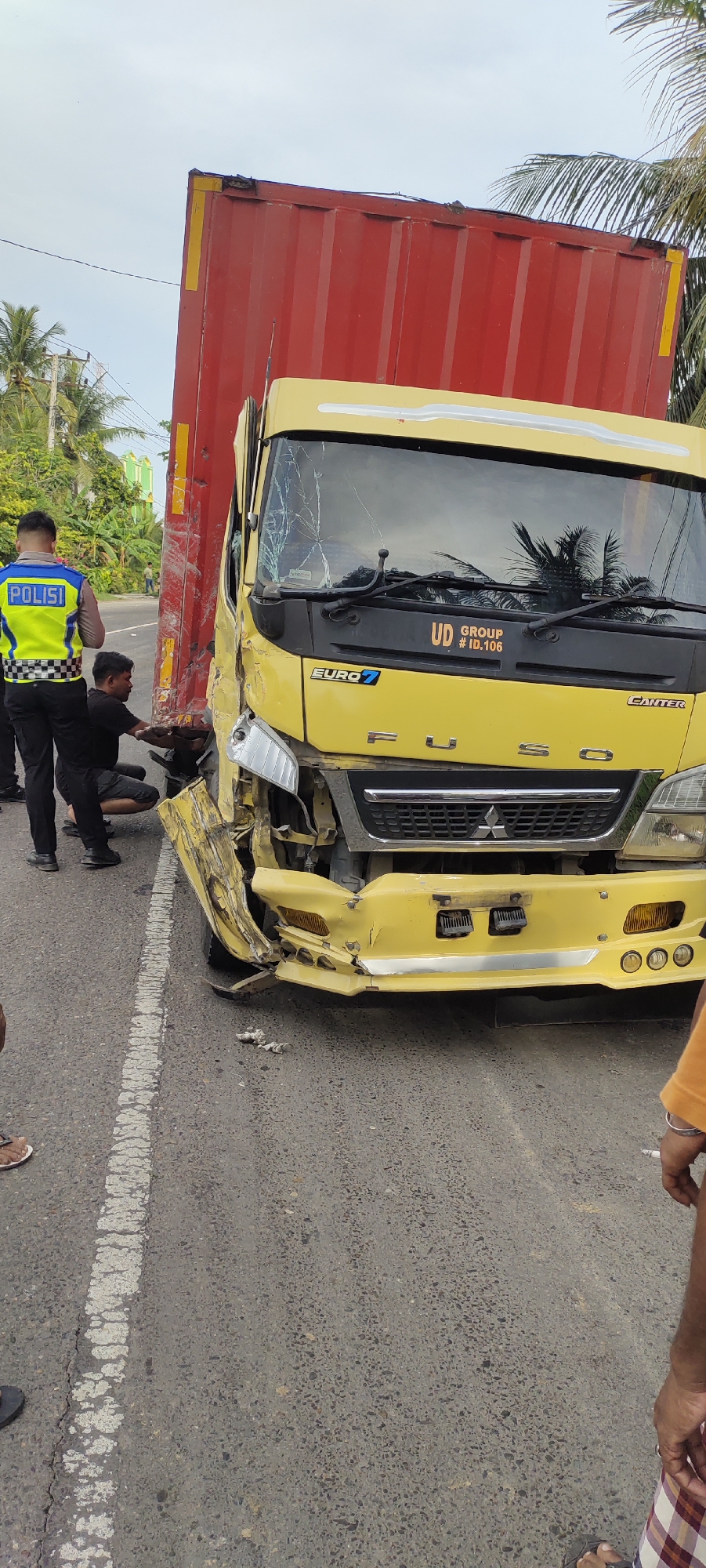 The image shows the aftermath of a traffic accident involving a yellow Fuso Canter truck with a red shipping container. The truck's front end is heavily damaged, with a shattered windshield and crumpled bumper and front quarter. A police officer in a blue and yellow uniform stands near the truck, along with a civilian who is kneeling down by the truck's front wheel. A man is seen on the right of the photo and another is partially cut off on the left.
The accident appears to have occurred on a paved road. Trees and foliage are visible in the background. Based on prior geographical knowledge, the location is likely Peurupok, Indonesia.