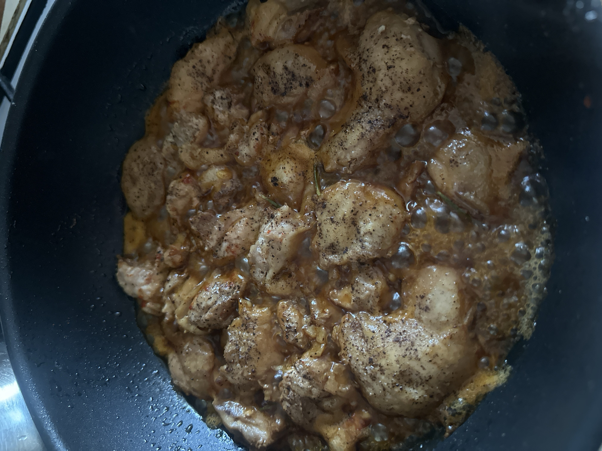 The image shows pieces of chicken being cooked in a dark non-stick frying pan. The chicken appears to be seasoned with black pepper and possibly paprika, and is simmering in a rich, golden-brown sauce. The pan is partially filled with the sauce, and small bubbles are visible around the chicken, indicating it is actively cooking. The background is out of focus and shows the edge of what appears to be a stovetop or kitchen counter. There is no indication of a specific location in the city of Peremyshliany, Ukraine.