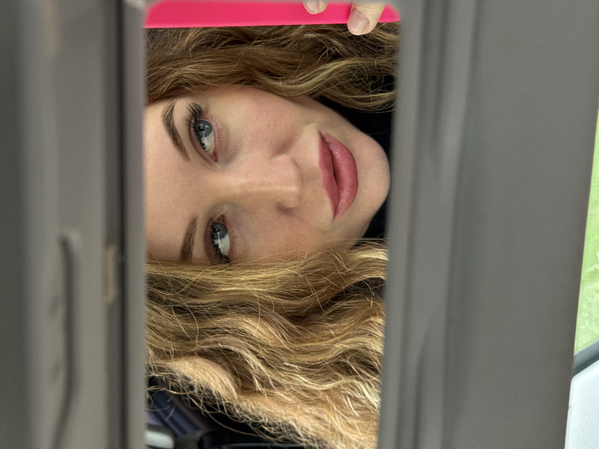 A woman with light skin, blue eyes, and blonde wavy hair is looking up into a partially open compartment. She has pink lipstick on and is wearing a dark-colored top. Above her is a pink object held by a hand. The scene is framed by gray metal or plastic structures. Outside the compartment, green and white objects are visible. The location is Chaikovsky, Russia.