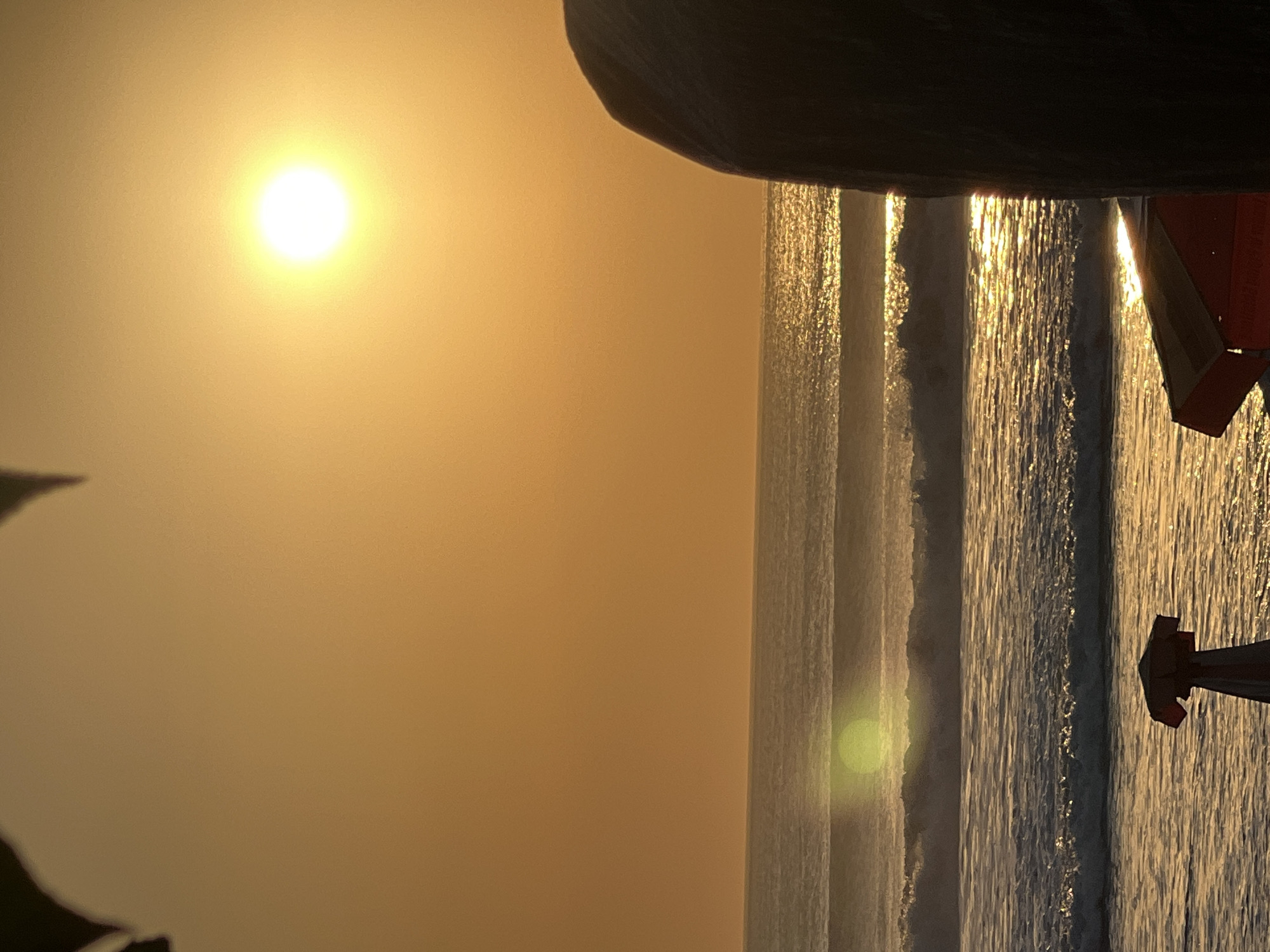 A sunset is captured over the ocean in Kuta, Indonesia. The sun, a bright orb, is positioned in the upper right quadrant of the frame, casting a warm, orange glow across the sky and water. The ocean exhibits distinct wave patterns, with the crests of the waves catching the sunlight and creating shimmering highlights. In the foreground, on the right side of the image, a large, dark, rounded object, possibly a rock or a piece of equipment, partially obscures the view. On the left side of the foreground, a portion of a beach umbrella and its supporting structure are visible, suggesting a beach setting. The overall atmosphere is serene and tranquil, indicative of a coastal environment at dusk.