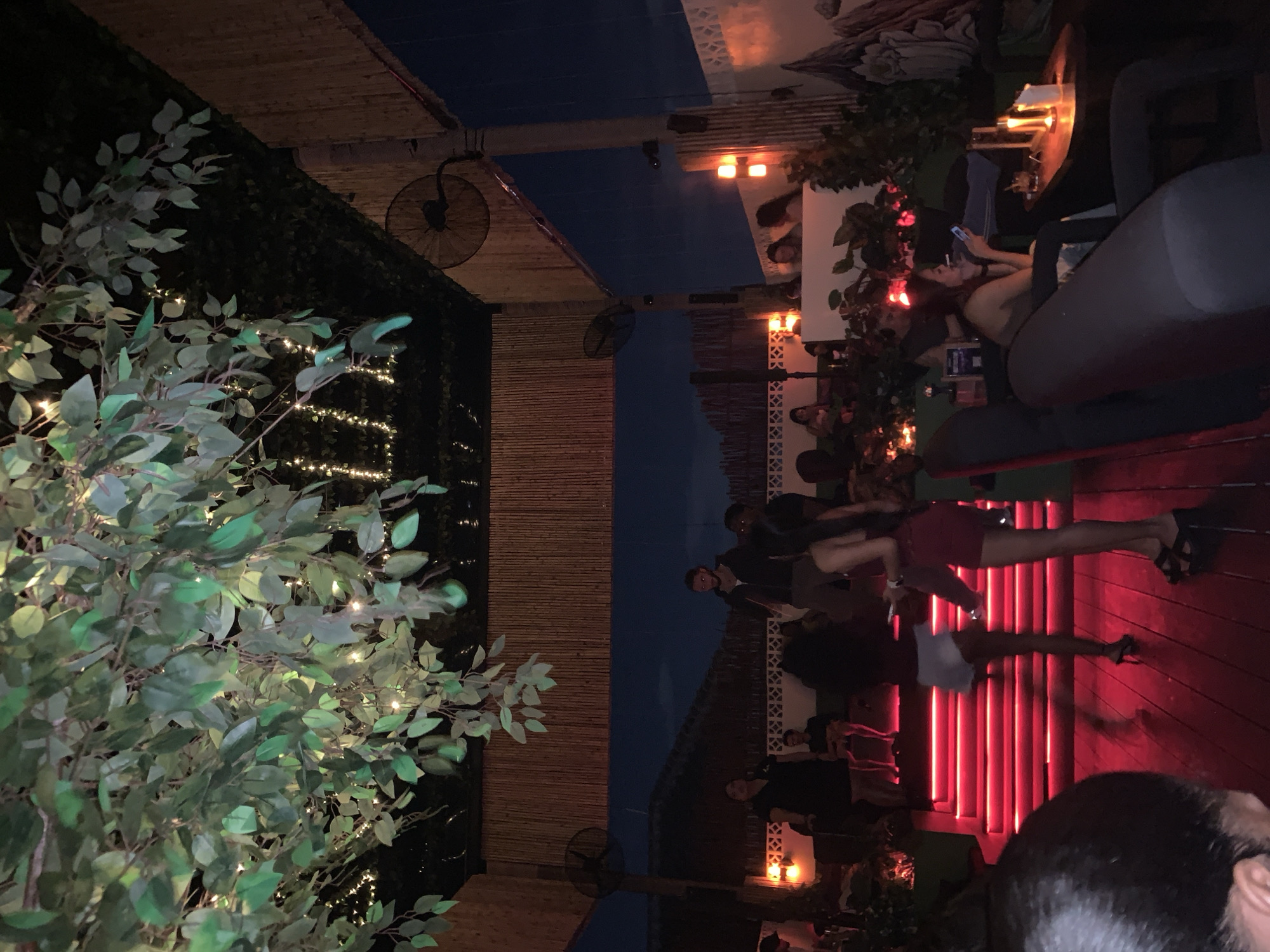 The image, rotated clockwise, captures an atmospheric evening scene at what appears to be an outdoor or semi-outdoor bar or lounge, consistent with a tropical resort setting in North Kuta, Indonesia. The time is clearly night, indicated by the dark sky and various sources of artificial light.
A prominent activity is taking place on a red-lit platform or stage in the mid-ground, where a woman in a red outfit is standing, seemingly performing or dancing. Several people are visible, forming an audience or guests of the venue. One woman is seated on a dark gray sofa to the right, engaged with her mobile phone and possibly smoking, with small, warm-toned lights on a nearby table. Other individuals are partially visible in the background, seated at tables or standing.
The setting is characterized by lush green foliage, particularly on the left, which is adorned with numerous string lights, adding to the festive ambiance. The architecture includes elements like woven bamboo screens and decorative white patterned dividers. Ceiling fans are installed overhead. In the background, a swimming pool with dark blue water is visible, suggesting a club or resort environment. The overall mood is social and relaxed, with people enjoying the entertainment and each other's company. No legible text is visible in the image.