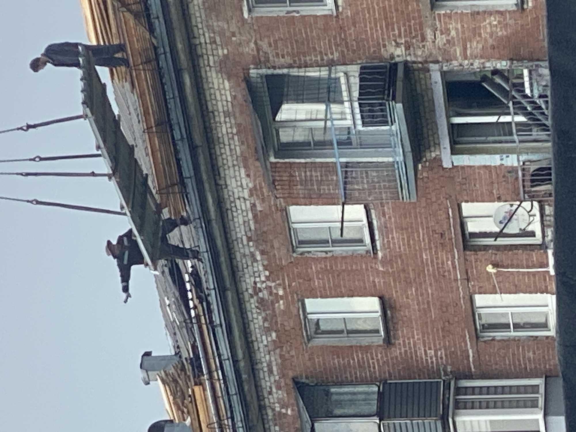 A group of construction workers are on the roof of a brick apartment building in Novokuznetsk, Russia, performing repairs or renovations. Two workers are visible on the roof. One worker, wearing a dark jacket and pants, is positioned on the far right, near a stack of wooden planks. He is looking down at the roof. Another worker, wearing a hat, dark jacket, and pants, is on the left side of the roof. He is holding his left arm out to the side, pointing towards the left, and appears to be directing or supervising the work. A large metal panel is being lifted by a crane, suspended by cables from above, and is positioned over the roof, between the two workers. The building has multiple windows and balconies, some with metal railings. The roof itself appears to be in a state of disrepair, with exposed wooden beams and some debris. The sky is overcast.