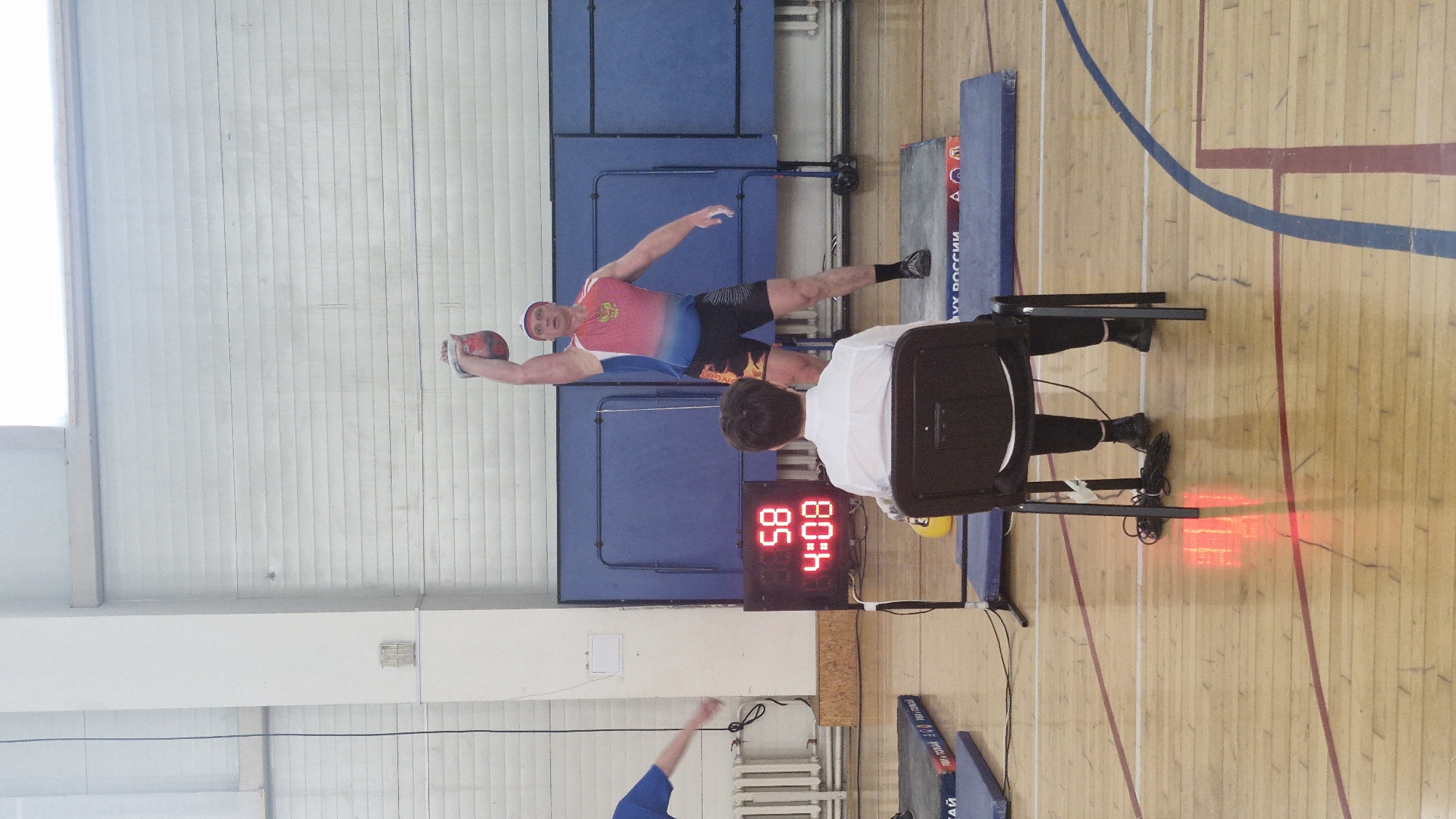 The image shows a man in a sports uniform holding a medicine ball in an indoor sports facility. The uniform is predominantly blue, white, and red, possibly indicative of the Russian flag. He is in a stance suggesting he is about to throw or has just thrown the ball. To his right, a person sits in a chair, seemingly observing or officiating. A digital timer displays the time "4:08:85." The setting appears to be a gymnasium with wooden floors marked with court lines. The background features a blue wall with panels. Based on the location given (Лобаново, Russia), this event is likely occurring within a sports facility in that city.