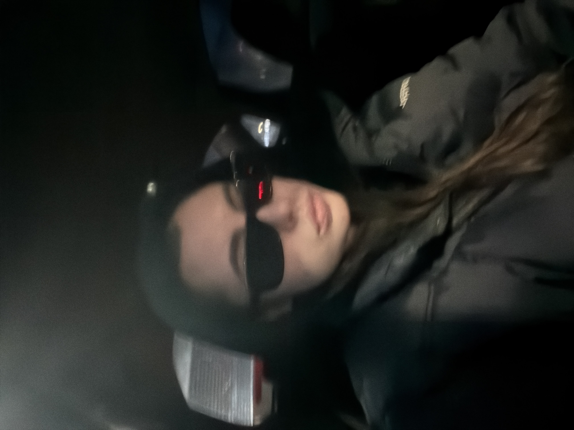 The media file consists of an original image and several crops, predominantly featuring a young person, likely female, inside a vehicle at night.
The central subject is a person with dark hair, seen from the chest up, captured in a diagonal orientation. They are wearing dark, rectangular sunglasses and a dark, possibly black, puffer jacket. Their lips are slightly parted, suggesting a relaxed or subtly playful expression.
The setting appears to be the dark interior of a vehicle, indicated by very low ambient light and specific illuminated details in the crops. Visible elements suggest car features: a red illuminated character, possibly an 'F' or a digit, and a lighter, C-shaped light are seen, likely originating from a dashboard or gear shift area. A reflective, rectangular object is partially visible beneath the person's chin, which could be a phone or another reflective surface.
The overall darkness of the image strongly implies it was taken at night. There is an illegible white logo or text on the left chest area of the jacket. The person seems to be posing for the camera, possibly a selfie. No specific location cues are visible within the image to suggest "Chaikovsky, Russia."
