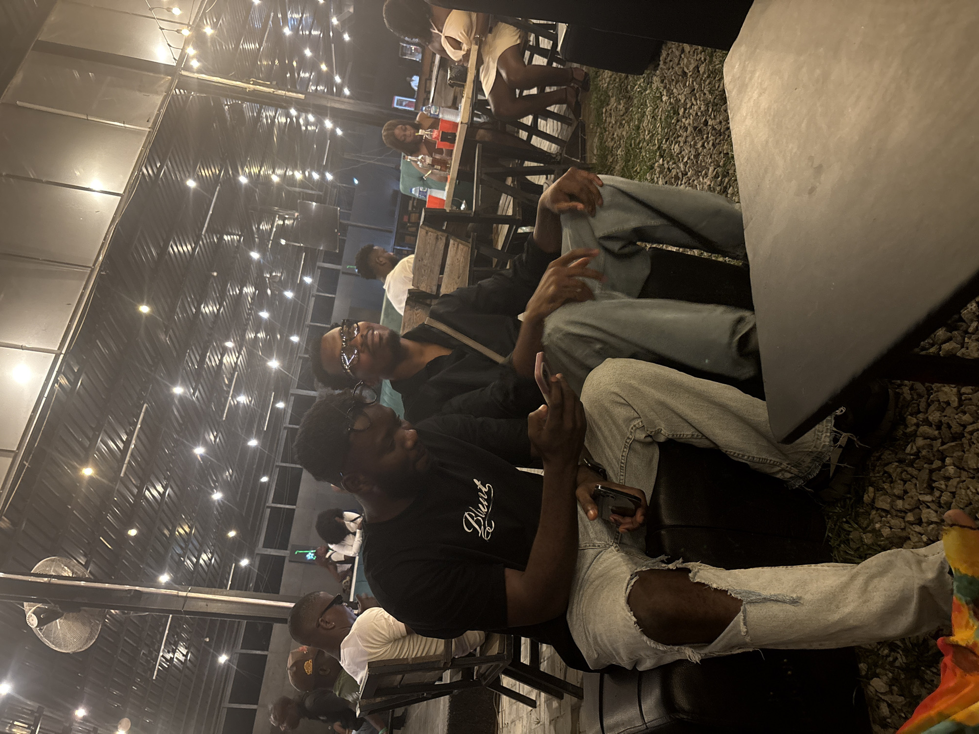 The image depicts a nighttime scene at an outdoor bar or lounge in Abuja, Nigeria. Several people are visible, seated at tables and engaged in conversation or looking at their phones. In the foreground, two men are prominently featured: one wearing a black t-shirt with white lettering and ripped jeans, and the other wearing a black shirt, glasses, and blue jeans. Both are looking at their phones. Other patrons are visible in the background, some at tables with drinks. The environment is illuminated by string lights suspended overhead, suggesting a relaxed and social atmosphere. The ground appears to be covered in gravel or small stones. A large fan is mounted on a pole.