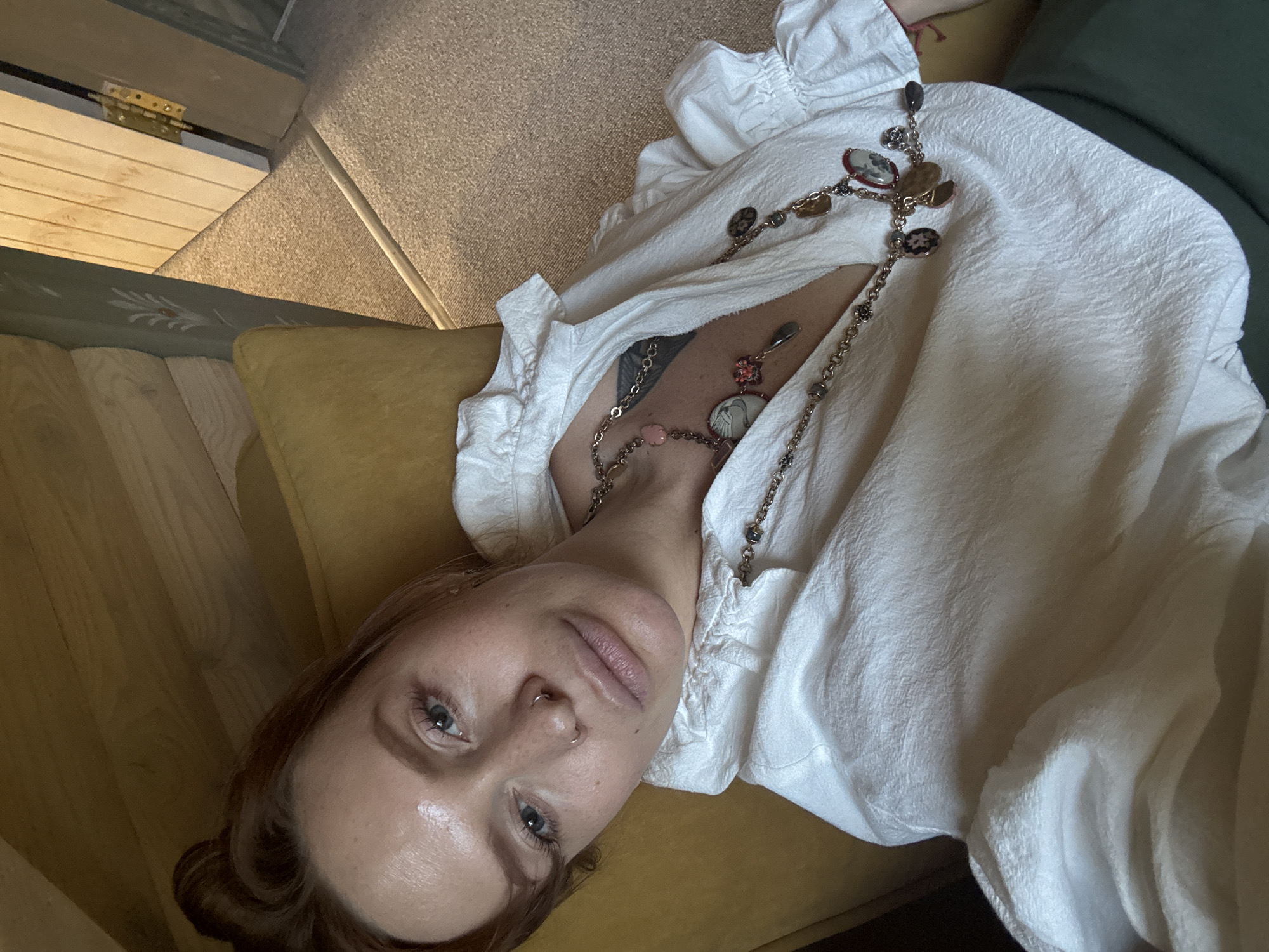 A woman with auburn hair, pulled up into a bun, lies on a yellow cushioned chair. She is wearing a white, ruffled shirt and a long, layered necklace with various charms. Her head is tilted, and she is looking directly at the camera. The background shows a portion of a room with wooden walls and a doorway leading to another room with carpeted flooring. The lighting suggests it is daytime. There are no identifiable landmarks to place this image specifically in the city of Suzdal, Russia.