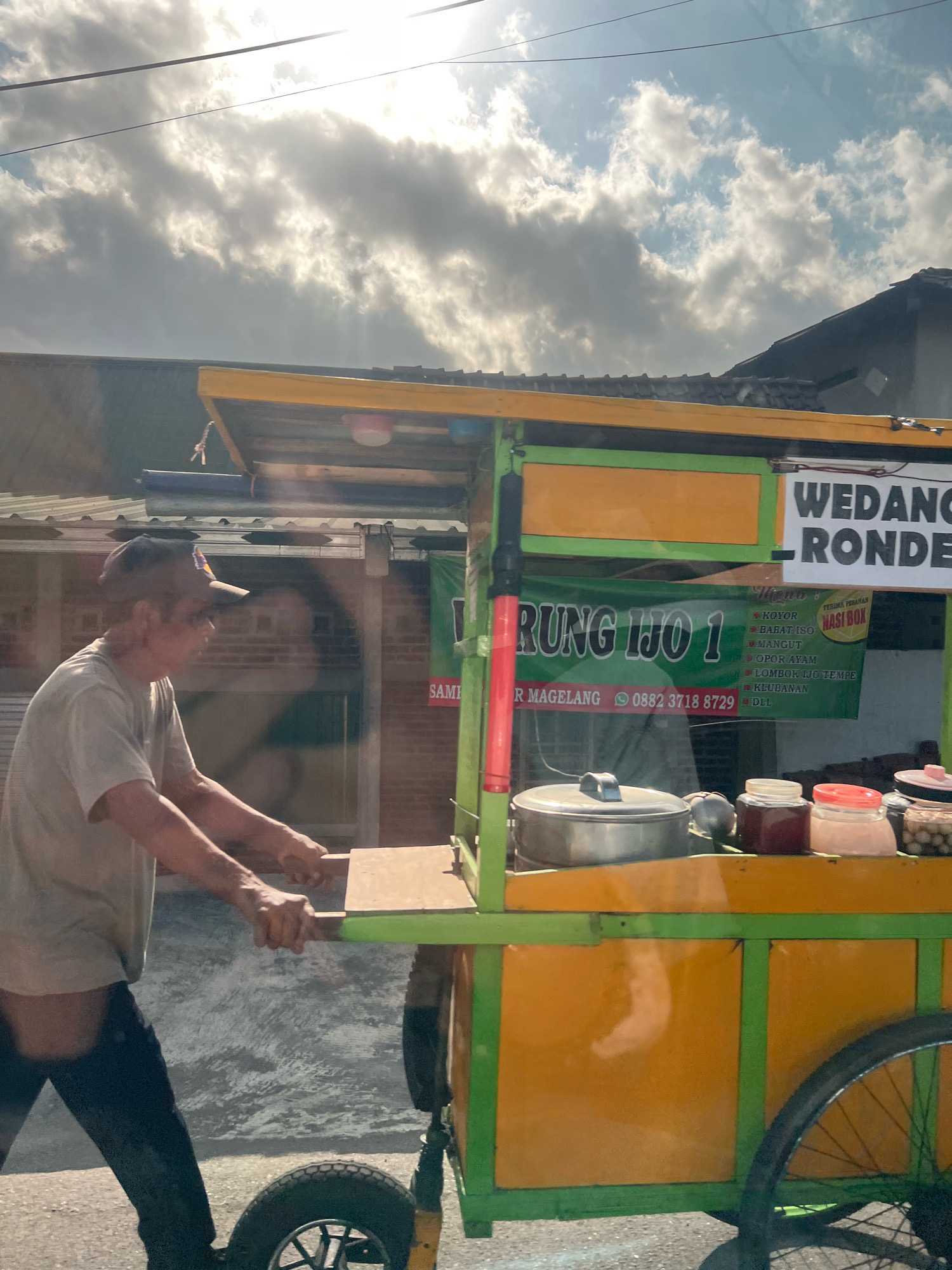 An outdoor, eye-level, full-shot photograph shows a man pushing a food cart in the city of Payaman, Indonesia. The sun shines brightly through the clouds in the sky. The man, who is wearing a light-colored t-shirt and dark pants, is in the process of pushing the cart forward with both hands. The food cart is bright yellow and green, with a selection of food items and jars visible on its surface. A sign on the cart reads "WEDANG RONDE". Another sign reads "WARUNG IJO 1" and lists various food items. The background consists of a brick building and a few houses. The street is paved.