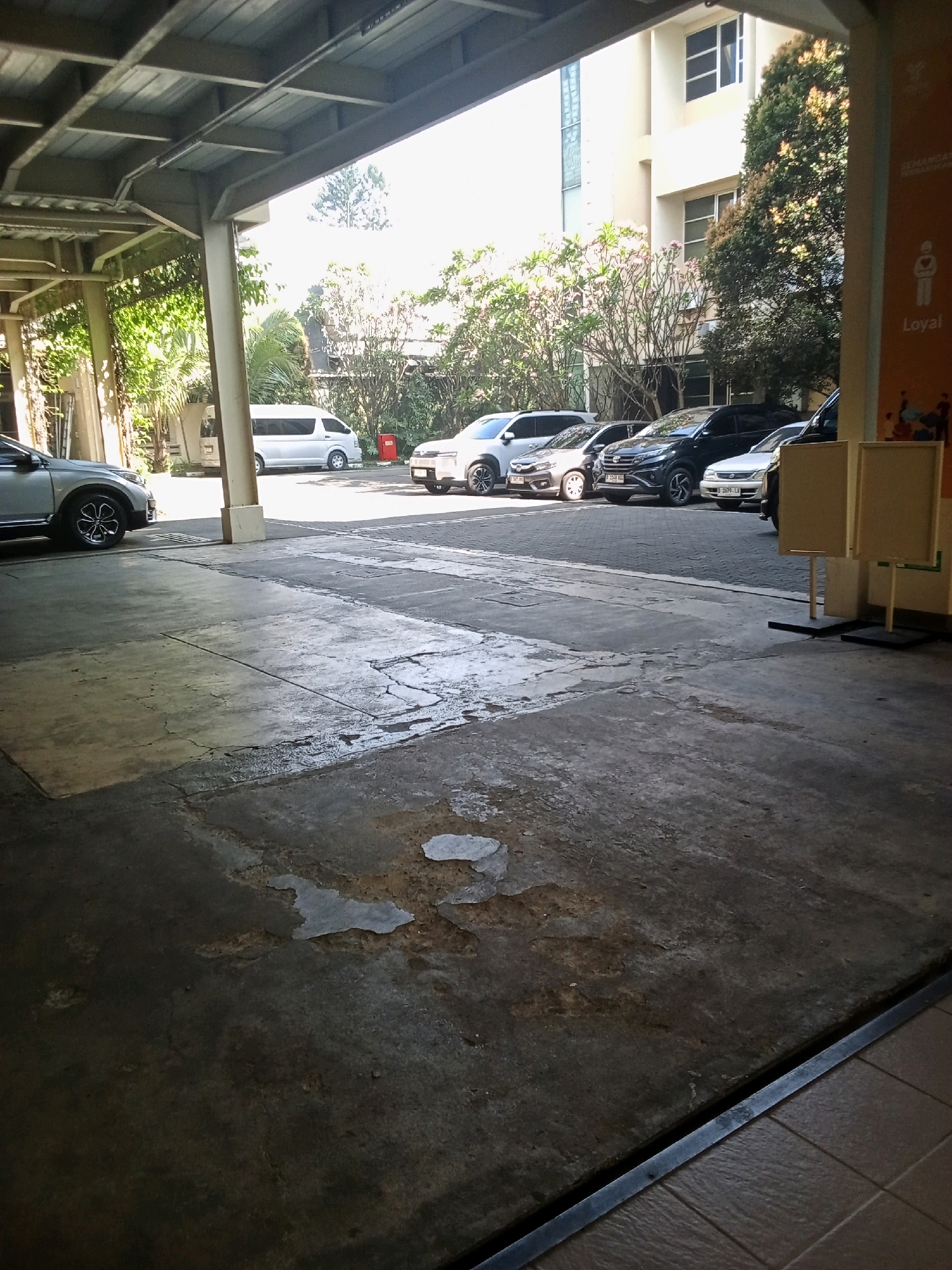 The image depicts a covered parking area, likely in an urban setting. The foreground shows a cracked concrete surface with a few damp patches, suggesting recent rain or a spill. In the middle ground, several cars are parked, including a silver SUV on the left, a white van, and a row of sedans and SUVs of various colors (black and silver). The cars are parked on an asphalt surface. Behind the cars, there are trees and shrubbery, and a light-colored building with multiple windows is visible in the background. A section of an orange sign with text and a pictogram of a person is present on the right side of the image. The overall impression is one of a functional outdoor space, possibly associated with a commercial building or residential complex. The presence of trees and the building suggests a developed area. The lighting indicates it is daytime, with bright sunlight filtering through the covered structure. The image does not provide specific clues about the exact location within Bandung City, Indonesia, beyond a general urban context.