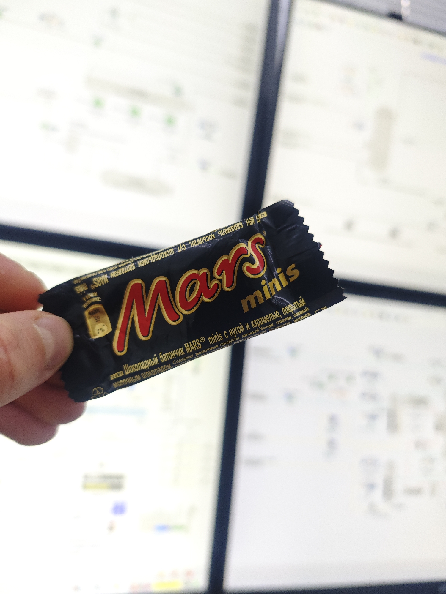 A hand holds a black "Mars minis" chocolate bar wrapper in front of several computer monitors displaying what appears to be code or data. The wrapper has red lettering for "Mars" and smaller gold lettering for "minis". Nutritional information is printed on the wrapper in a smaller font. The background consists of the blurry displays of the computer monitors.