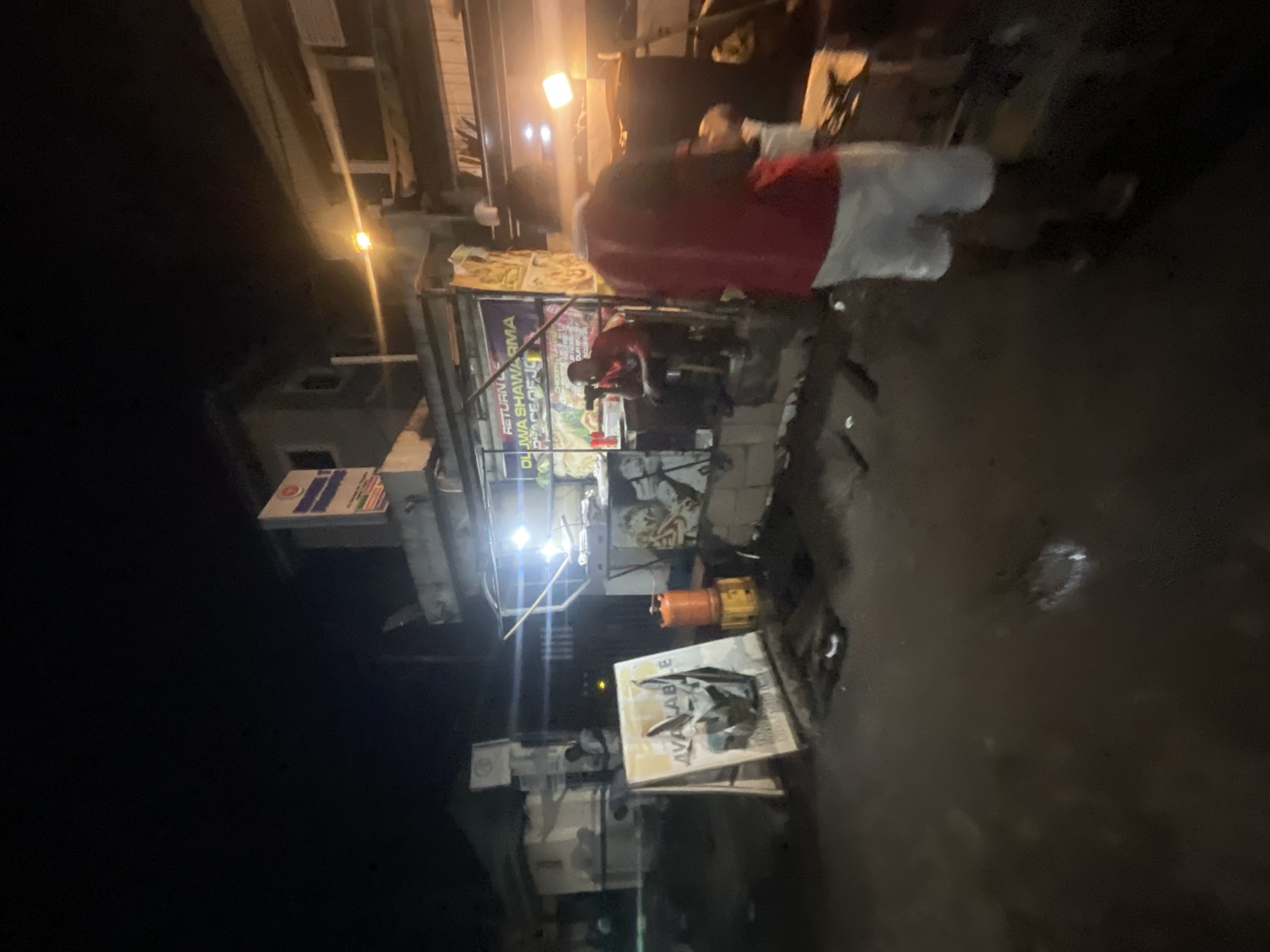 This is a nighttime photograph of a street scene in Akowonjo, Nigeria. In the foreground, a person wearing a red shirt and white shorts is walking away from the camera. To the left, an A-frame sign displays an image and the word "AVAILABLE". Further back, a food stall is illuminated by bright lights, with several people visible. One person is seated at the stall, and another is standing. A gas cylinder is positioned next to the stall. The environment appears to be a roadside with a paved surface, and there are buildings in the background.
