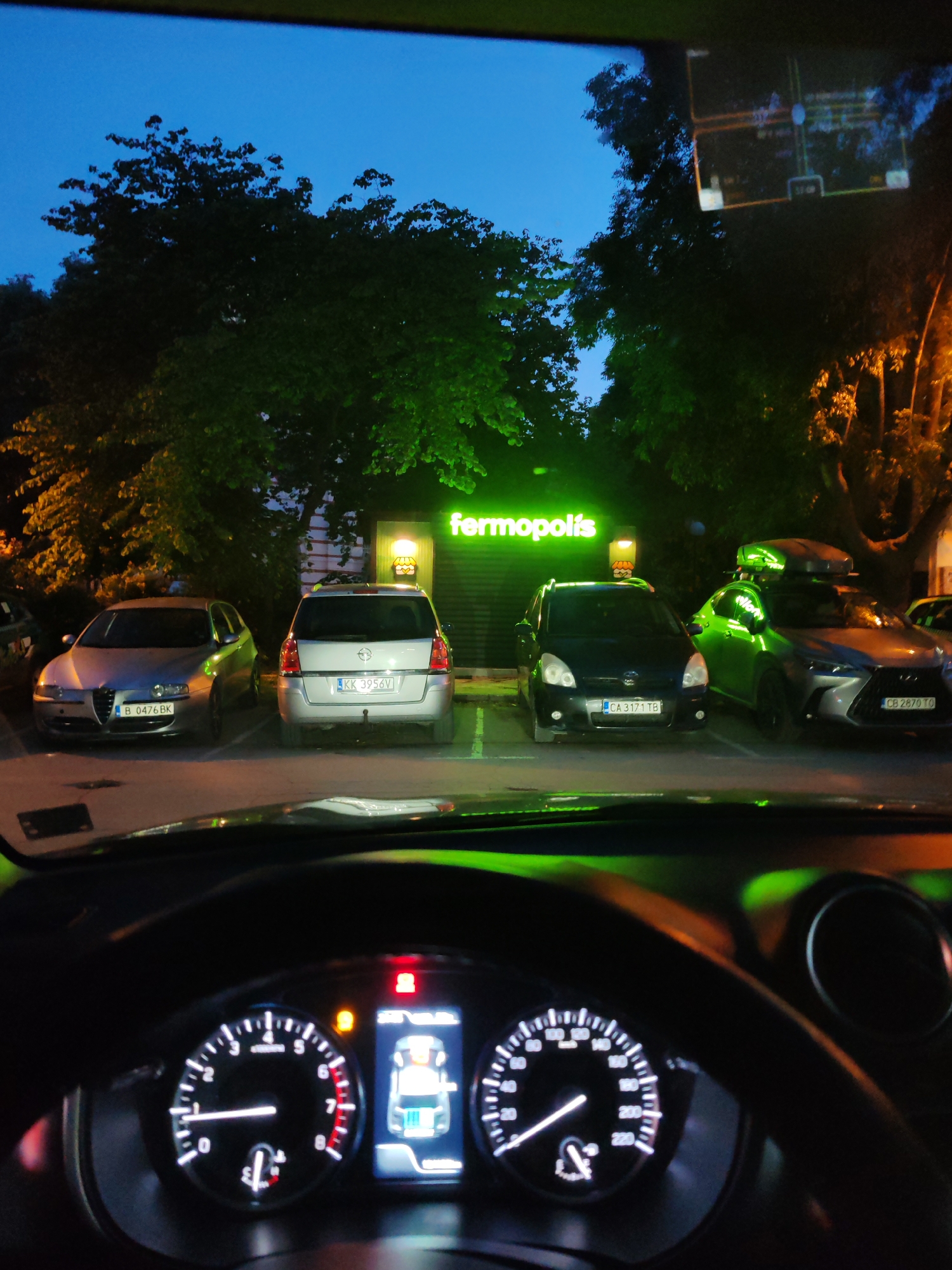 Captured from inside a car, the image reveals a nighttime scene in front of the "fermopolis" fast-food restaurant, recognizable by its illuminated green signage. Four cars are parked in front of the establishment. Reading from left to right, the first is a silver Alfa Romeo (license plate B 0476 BK), followed by a silver Opel Zafira (license plate KK 3956V), a black Toyota Corolla Verso (license plate CA 3171 TB), and a grey Lexus SUV with a roof cargo carrier (license plate CB 2870 TO). The sky is a deep blue transitioning to dark, and trees surround the restaurant.
Inside the car, the dashboard and steering wheel are visible, with illuminated gauges and warning lights. A navigation screen in the upper right reflects the same outdoor scene.