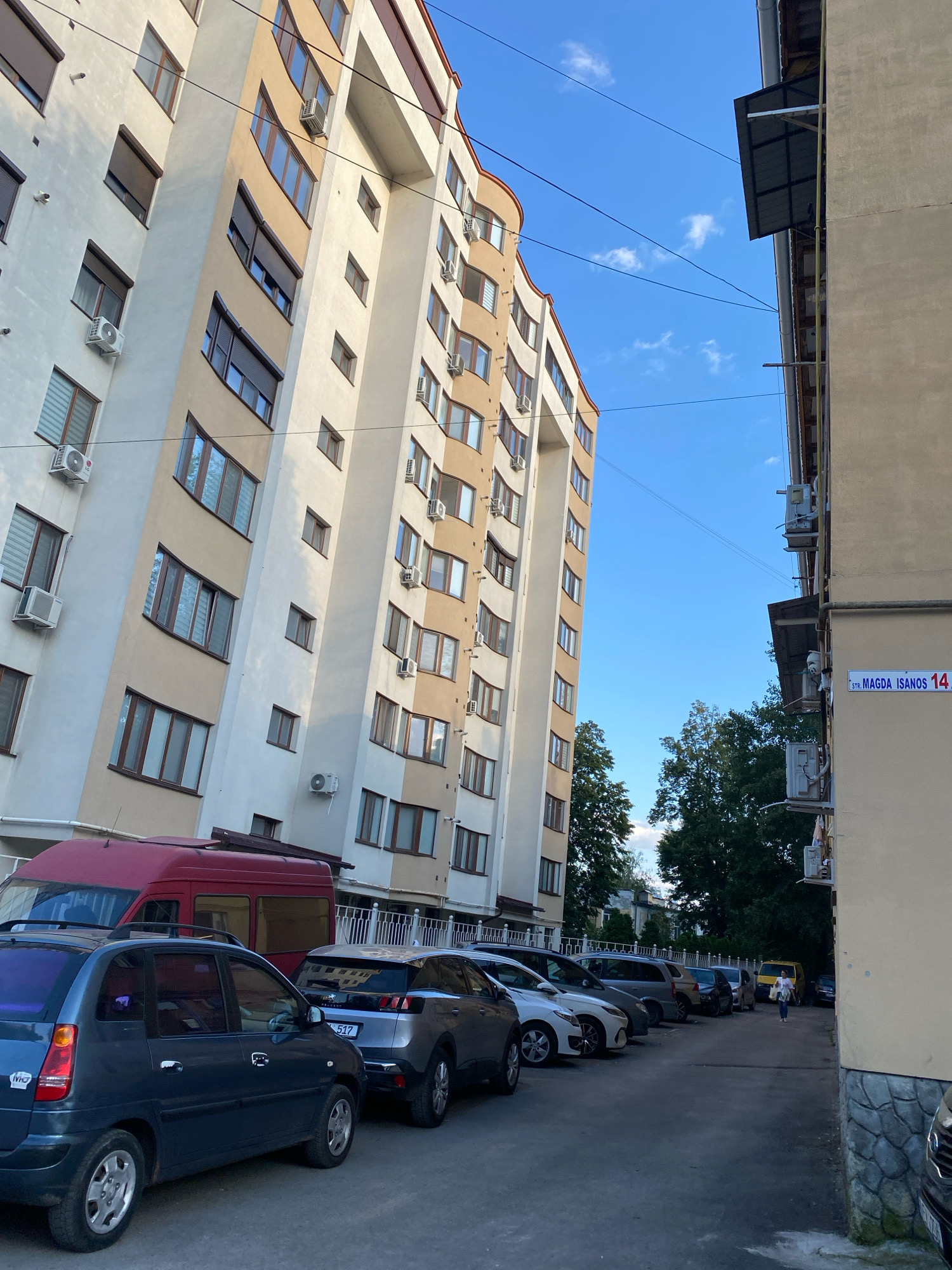 The image captures a street scene in Chișinău, Moldova, specifically on Strada Magda Isanos 14. A tall, modern apartment building with beige and white facade dominates the left side of the frame, angled towards the viewer. It features numerous windows, many equipped with roll-down shutters, and several air conditioning units are visible mounted on its exterior. Power lines crisscross the blue sky, which has a few wispy clouds. To the right, a smaller, light-colored building runs parallel to the apartment block, with its own visible air conditioning units and a gutter system. A street sign on this building clearly reads "STR. MAGDA ISANOS 14". In the foreground, a parking area is occupied by several vehicles. These include a dark blue hatchback on the far left, a red van behind it, and a row of other cars parked further down the lot, including a silver Peugeot SUV. A woman in white is walking away from the viewer towards the background on the right side of the parking lot. Lush green trees are visible in the background, partially obscuring other buildings. The overall impression is an urban residential environment under a clear sky.