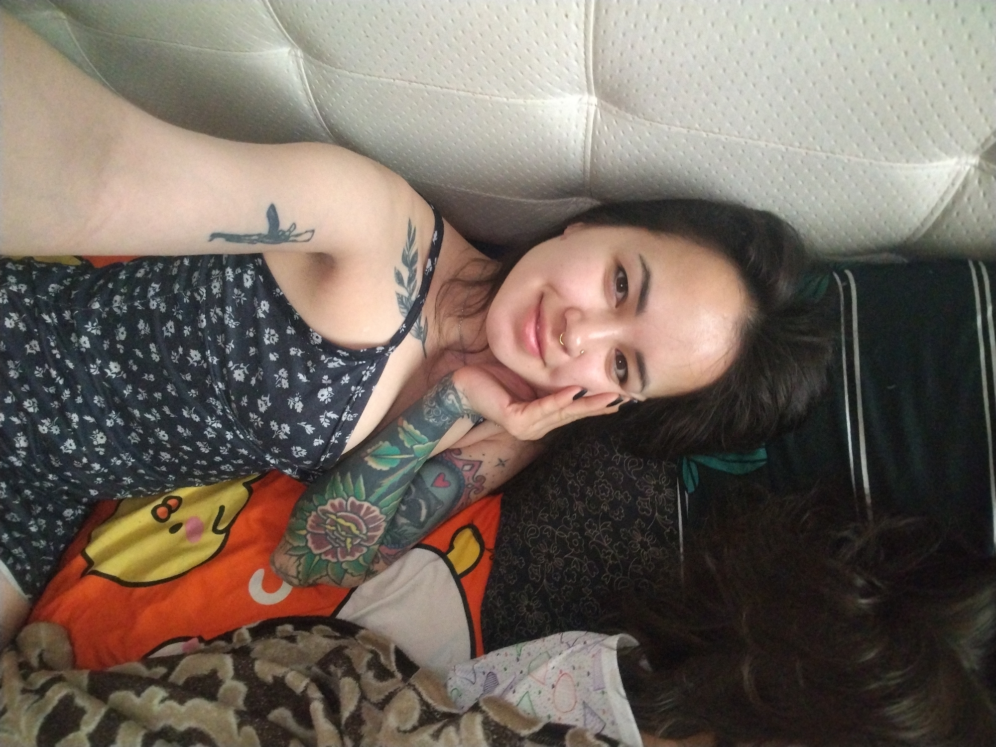 A young woman lies on her side on a bed. She rests her head on her hand, with her fingers near her cheek. She has dark hair and a nose ring. She wears a sleeveless dress with a floral pattern. Her left arm has several tattoos. She lies on a pillow with a cartoon face on it. A dark, striped cushion is behind her. A patterned blanket covers the lower portion of her body. The headboard of the bed is white. The location is unknown.