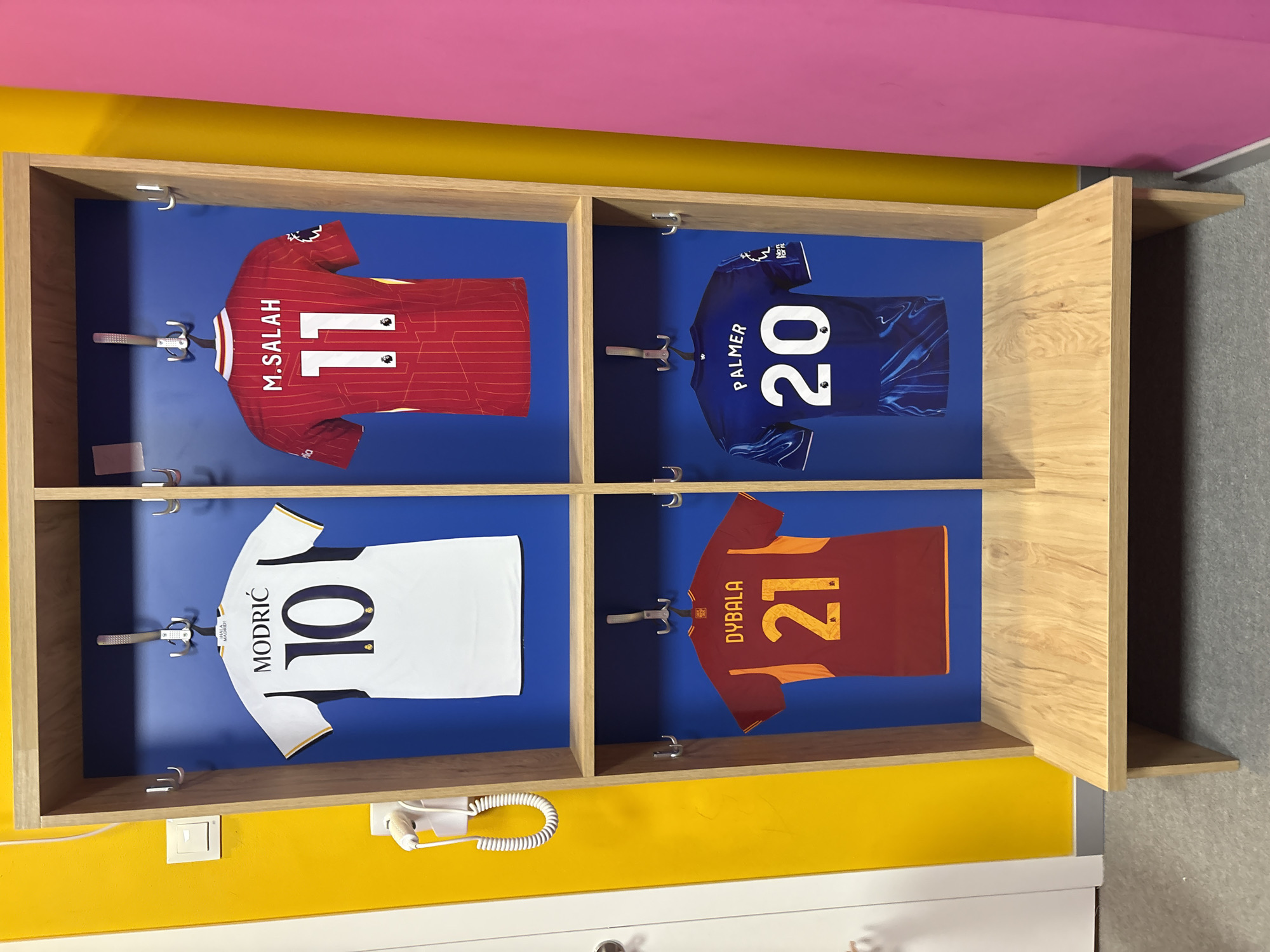 The image depicts a locker room scene featuring four soccer jerseys hanging in separate cubbies. The jerseys are displayed on hangers against a bright blue background. From left to right, top row: a white jersey with "MODRIĆ" and the number "10" in navy blue, and a red jersey with "M.SALAH" and the number "11" in white. Bottom row: a maroon and orange jersey with "DYBALA" and the number "21" in orange, and a blue jersey with "PALMER" and the number "20" in white. The locker unit is made of light-colored wood and is set against a bright yellow wall. The floor appears to be carpeted. The scene is devoid of any people or animals. The overall impression is a clean and organized display of sports memorabilia. This scene is not located in Valencia, Spain.